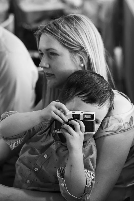 A woman holding a young boy who is taking a picture with a compact camera, in an indoor setting.