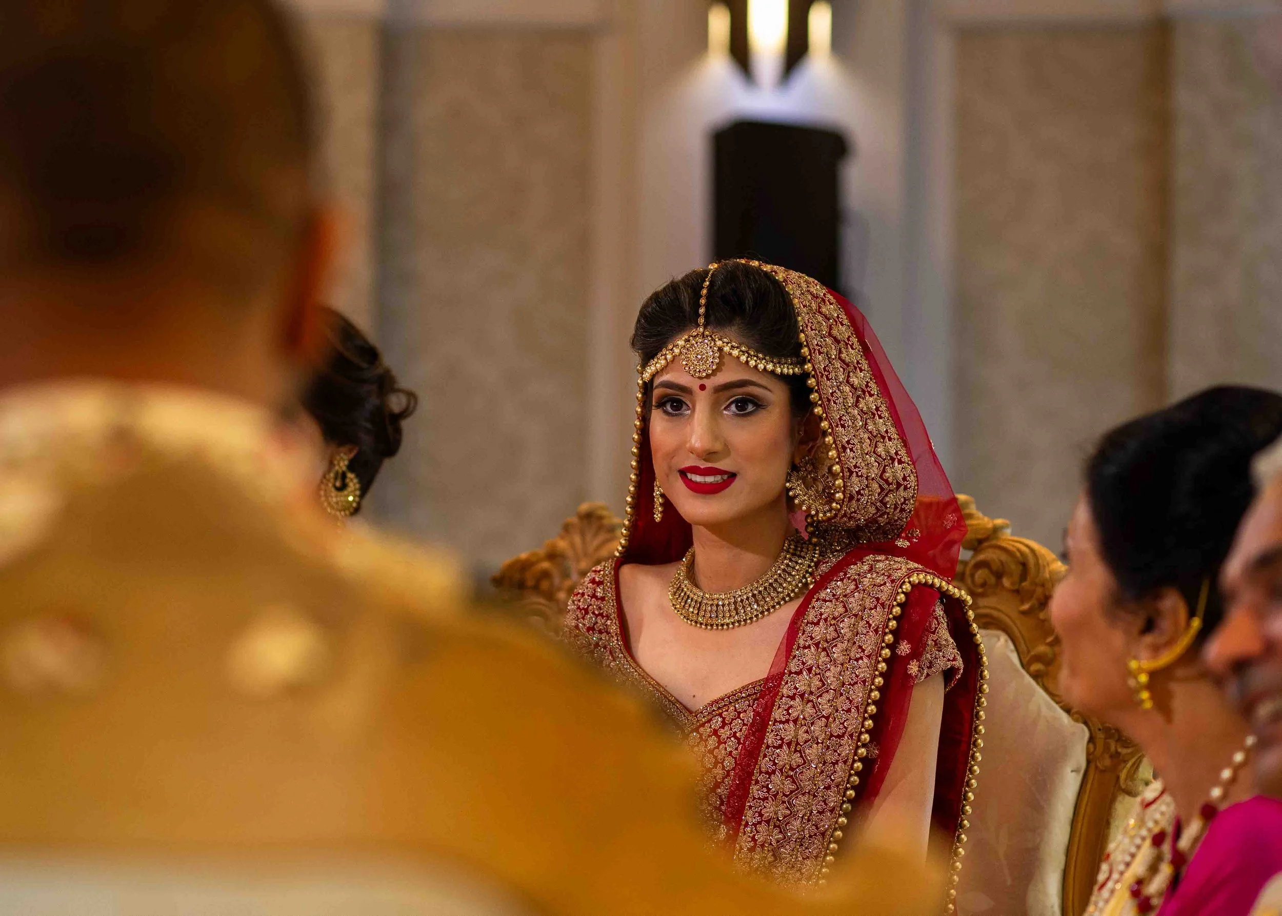A woman dressed in traditional Indian wedding attire, wearing a red and gold embroidered sari, jewelry including a forehead ornament, necklace, and earrings, sitting on a ornate chair, smiling.