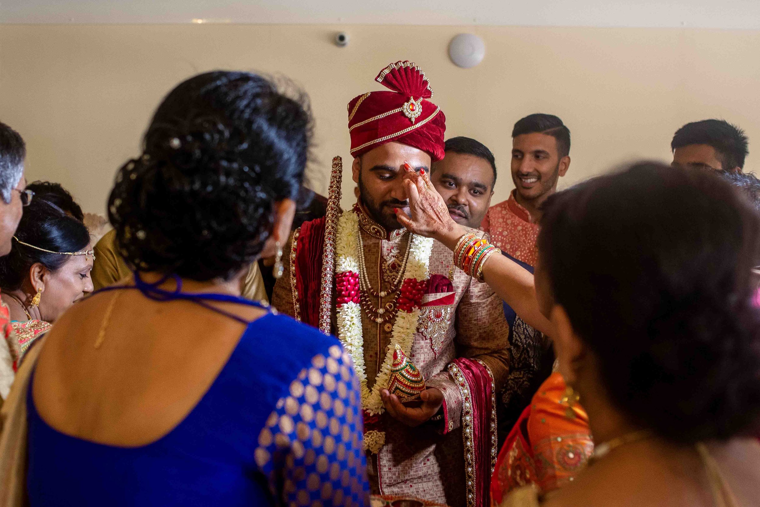 Indian groom dressed in traditional wedding attire, wearing a red turban and floral garlands, as female guests apply sindoor on his forehead during a wedding ceremony, surrounded by family and friends.