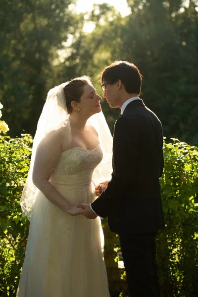 Bride and groom holding hands, about to kiss outdoors during sunset, lush greenery in the background.