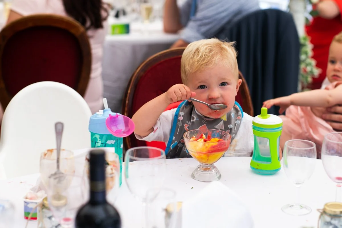 A young blonde boy with a bib eating dessert with a spoon at a birthday party or family gathering, surrounded by drinks and partyware on a table.