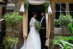 Bride and groom exchanging vows under a canopy at an outdoor wedding ceremony, surrounded by greenery and stone building.