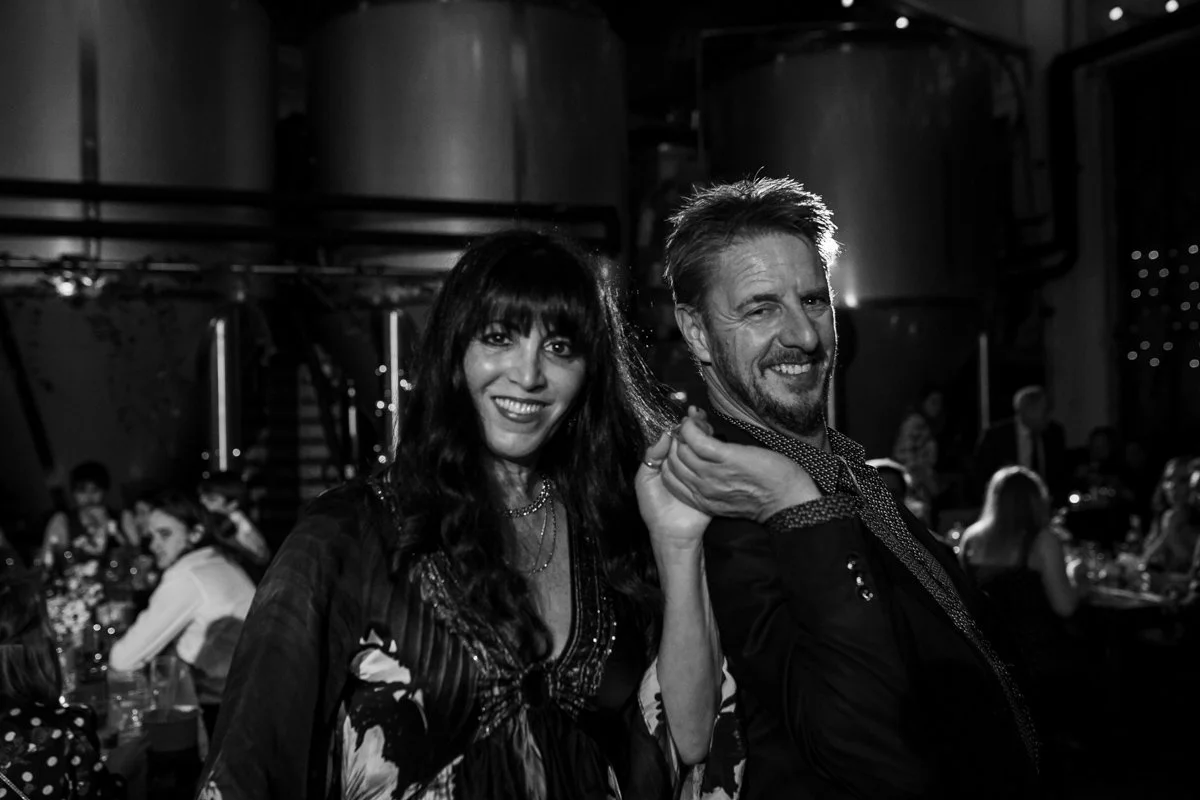 A black and white photo of a woman and a man dressed nicely at a social gathering or party. The woman has long dark hair and is smiling, while the man has short hair, a beard, and is winking.
