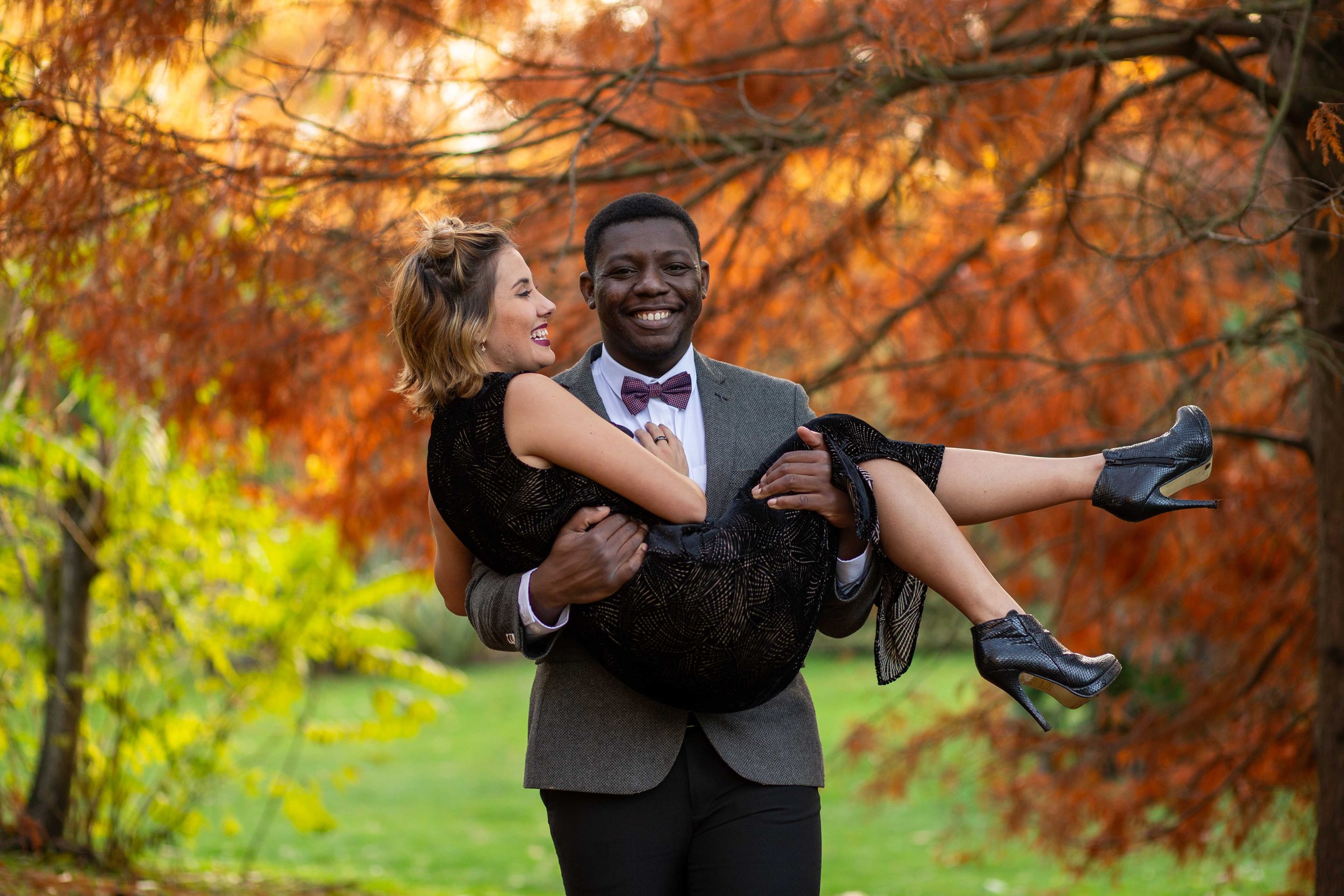 A man in a gray suit carrying a woman in a black dress in a park during autumn, with orange and yellow leaves on the trees.