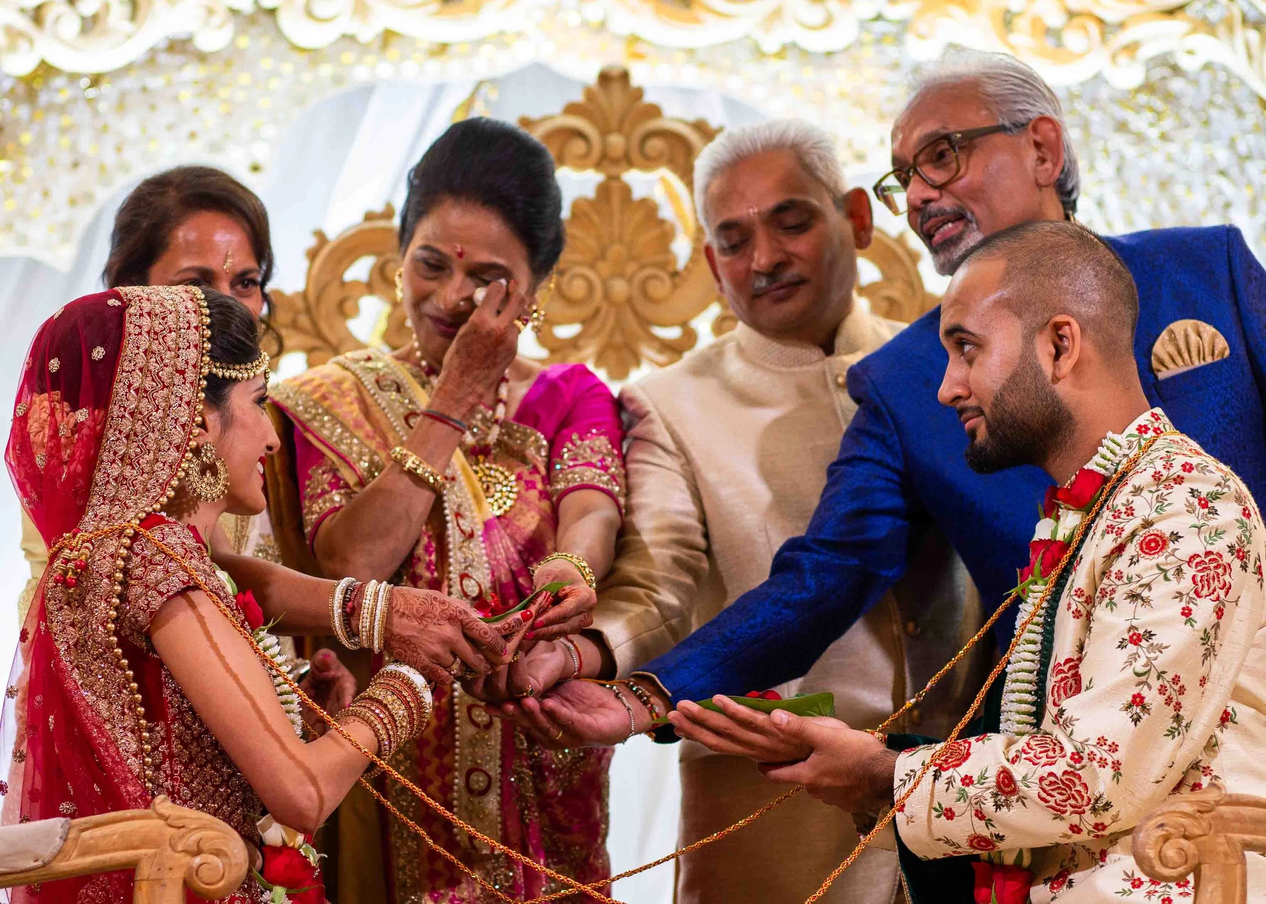 Indian wedding ceremony with bride and groom exchanging rings, surrounded by family members in traditional attire.