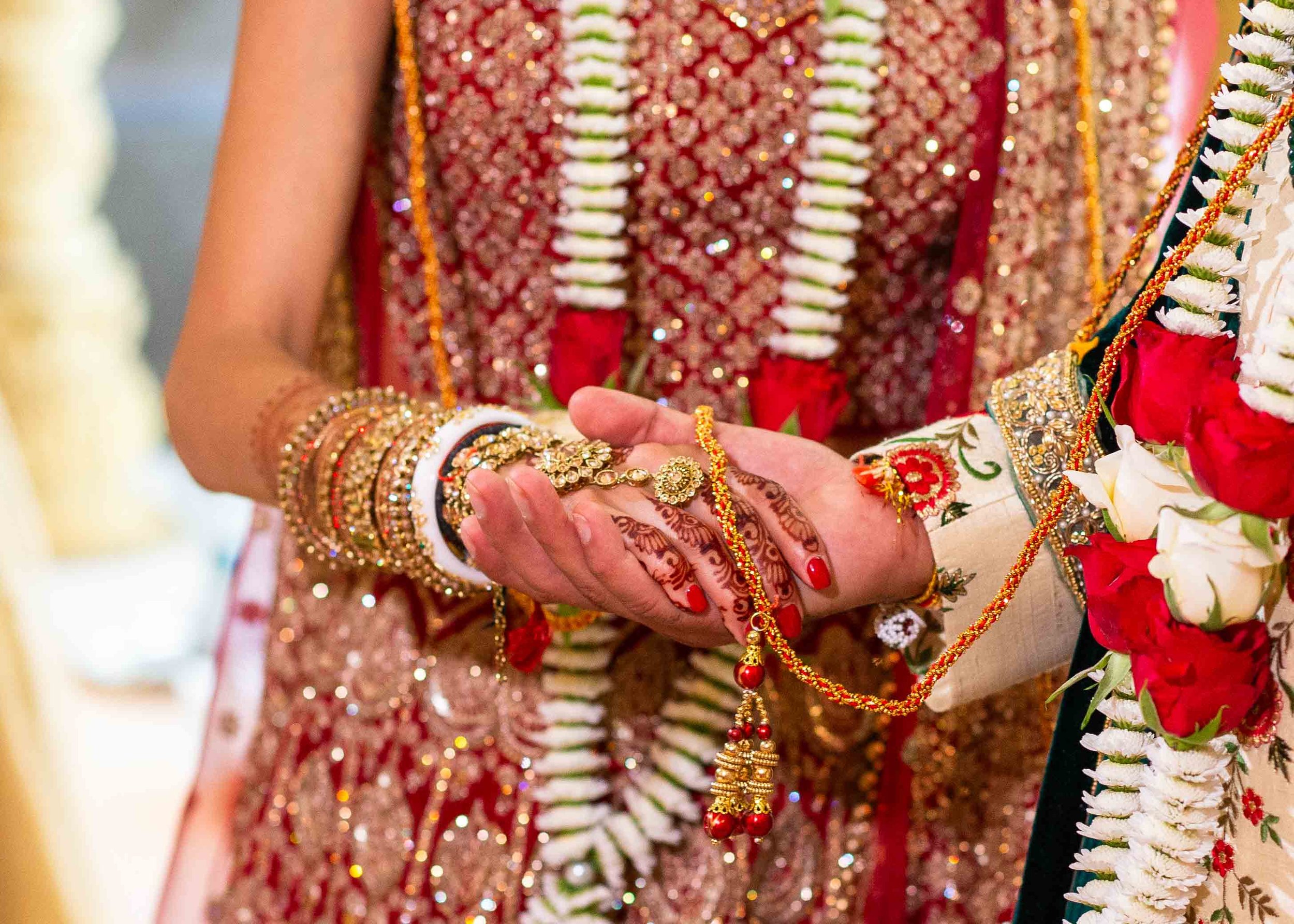 Close-up of a traditional Indian wedding ceremony showing hands adorned with jewelry and henna, exchanging floral garlands, with intricate clothing and floral decorations in the background.