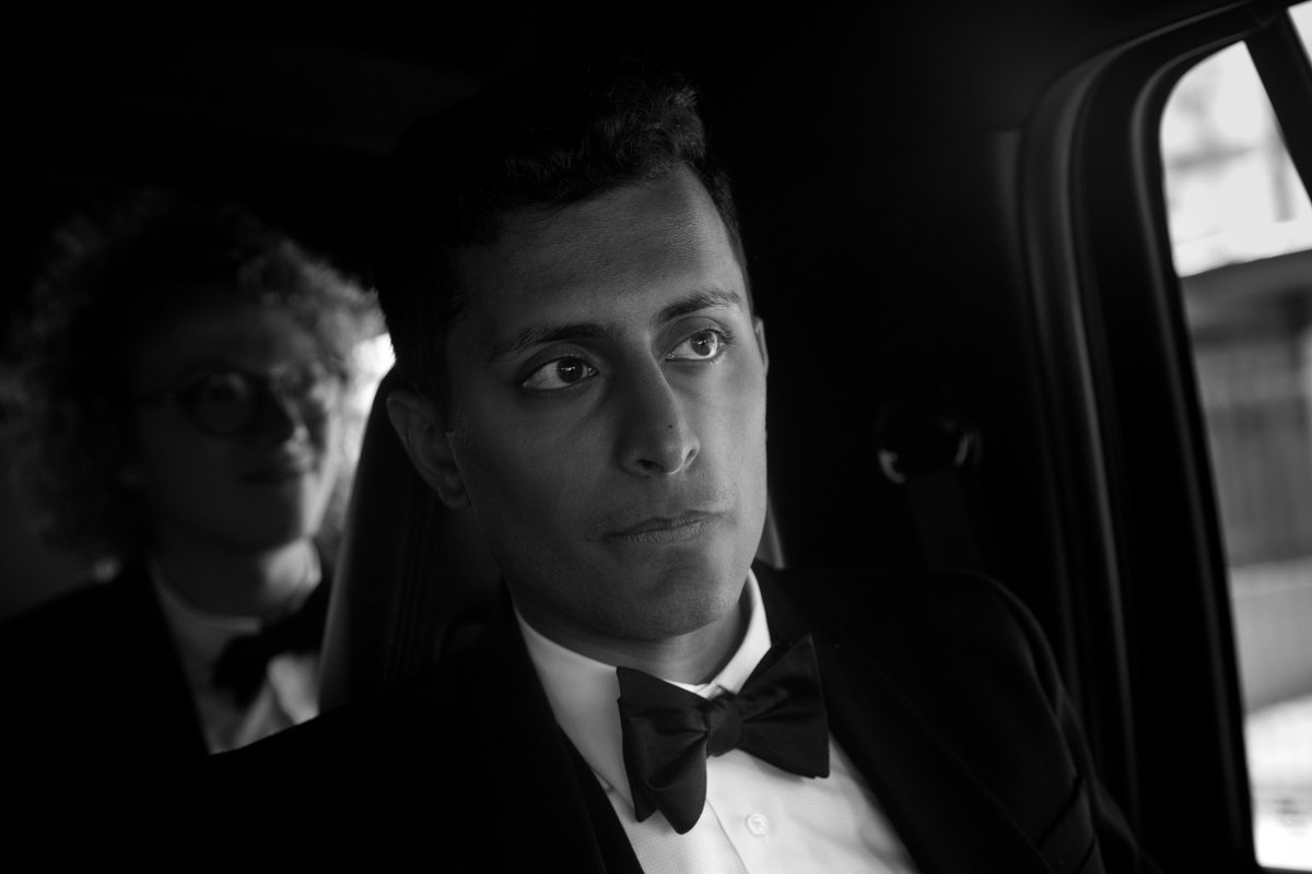 A black and white photo of two men in tuxedos seated inside a vehicle. One man is in the foreground with a serious expression, and another man with glasses is in the background, slightly out of focus.