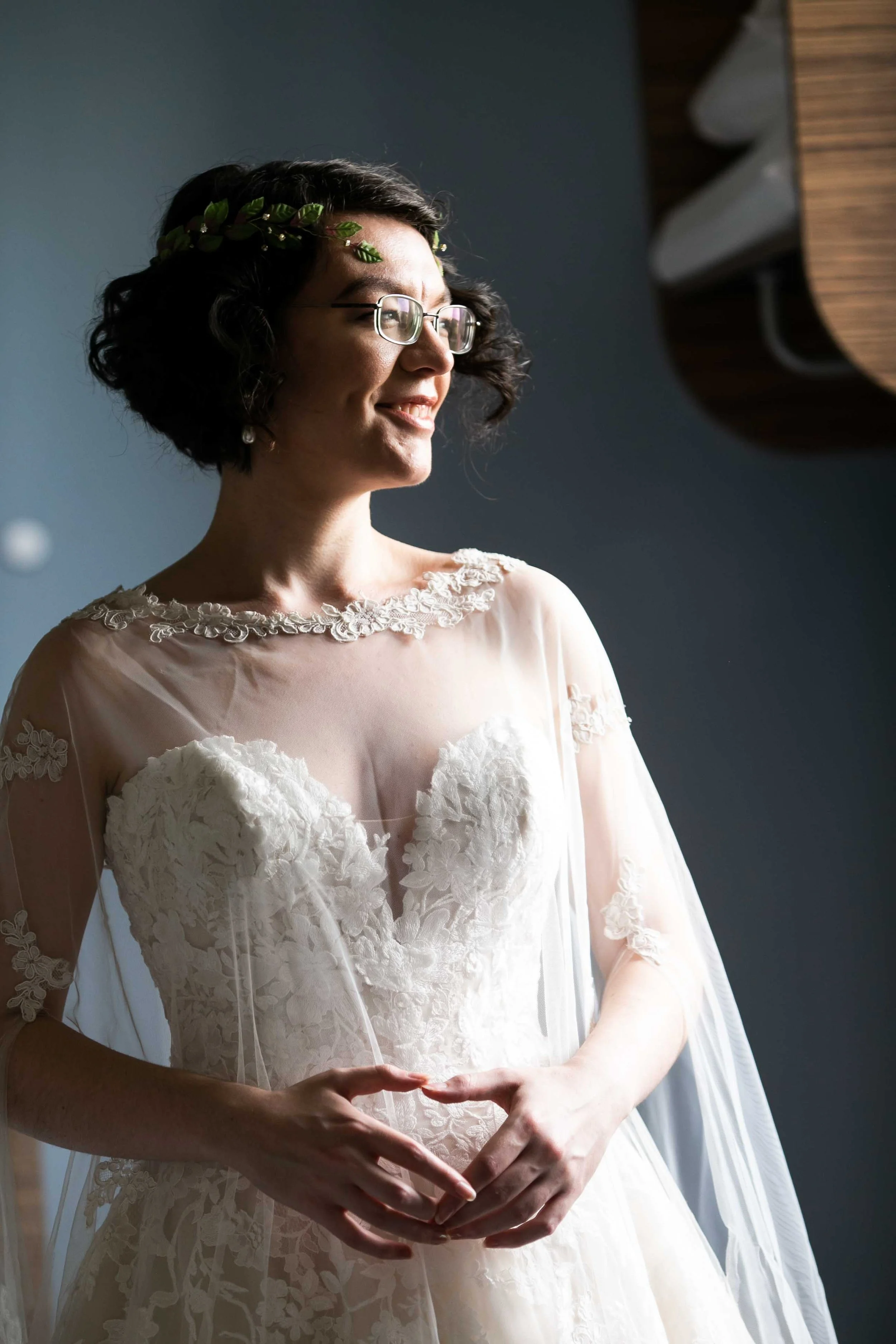 A woman in a wedding dress with lace details and sheer fabric, wearing glasses and a green leaf headpiece, standing near a window