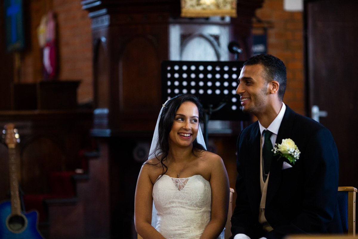 A bride and groom sitting side by side during their wedding ceremony, smiling at each other inside a church with wooden decor.