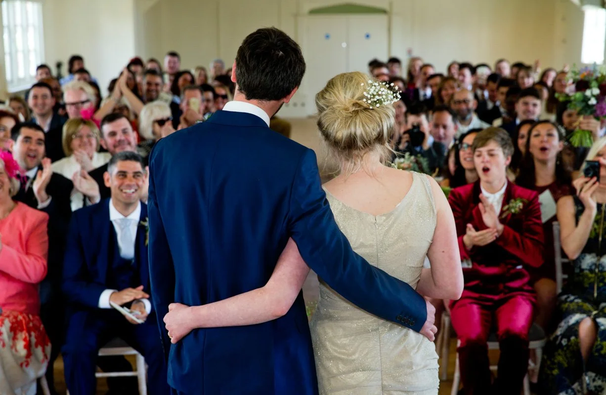 A bride and groom stand together, embraced from behind, facing a joyful wedding audience in a decorated indoor venue.