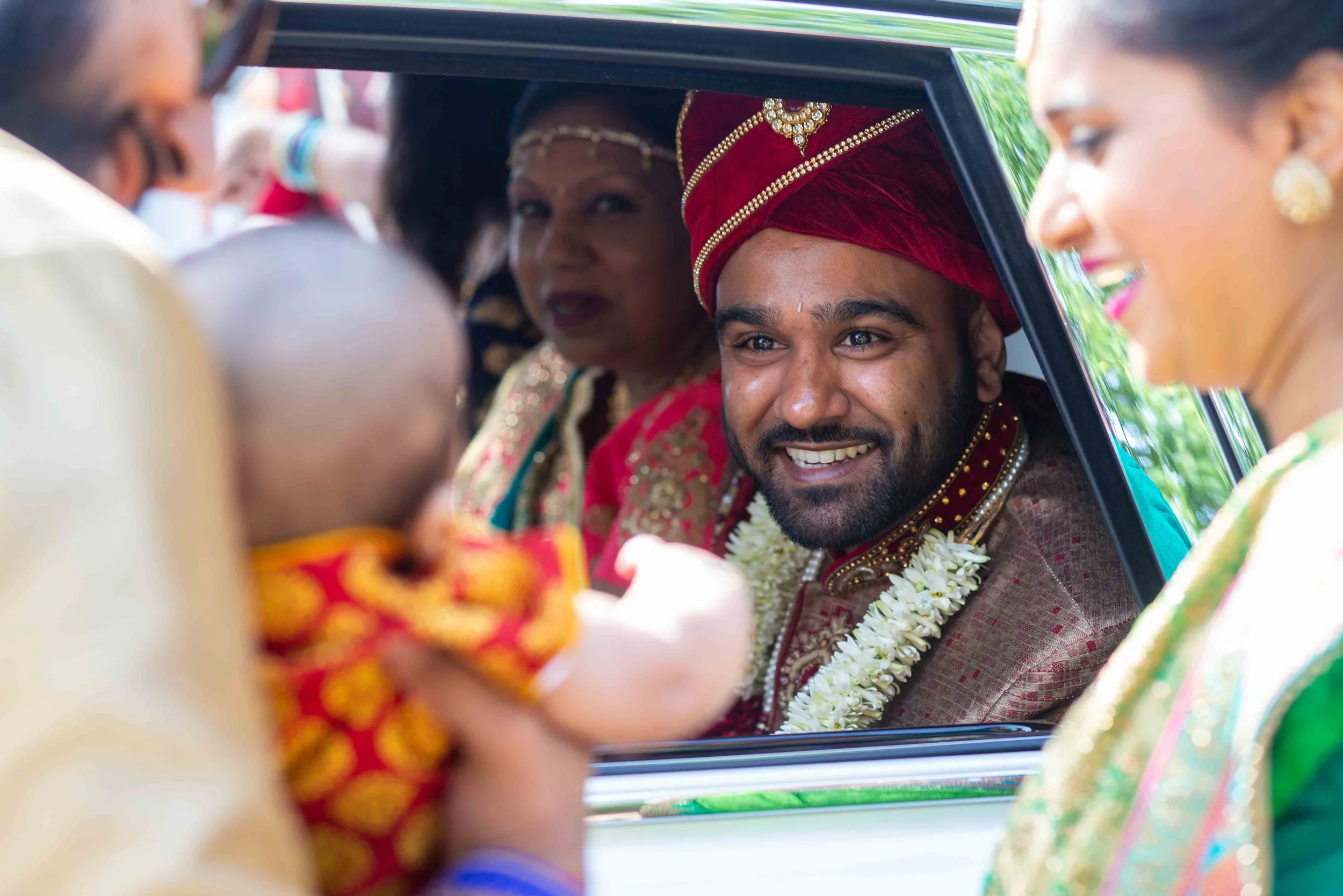 A groom in traditional Indian attire smiling as he sits in a car during a wedding, surrounded by family members in colorful traditional clothing.
