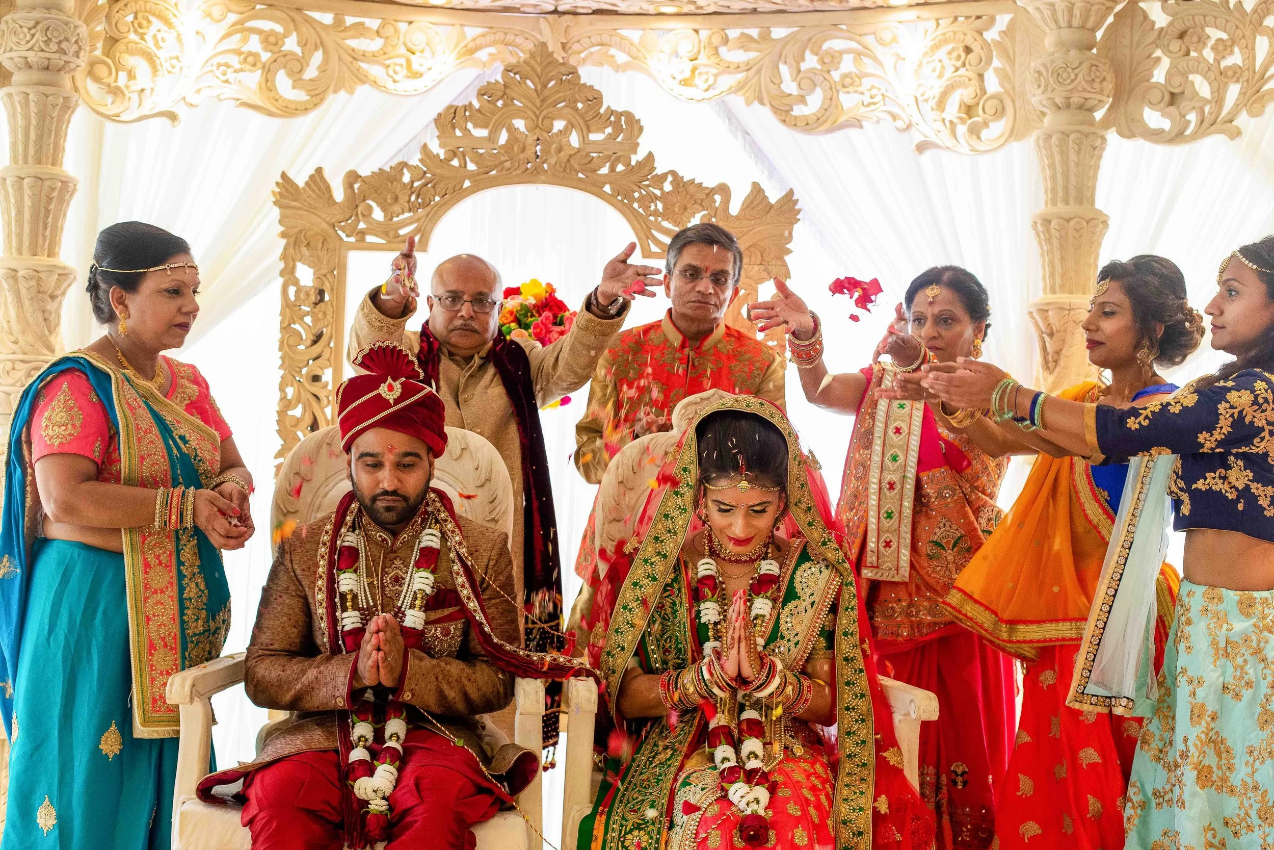 Indian wedding ceremony with a bride and groom seated in the center, surrounded by family members performing rituals.