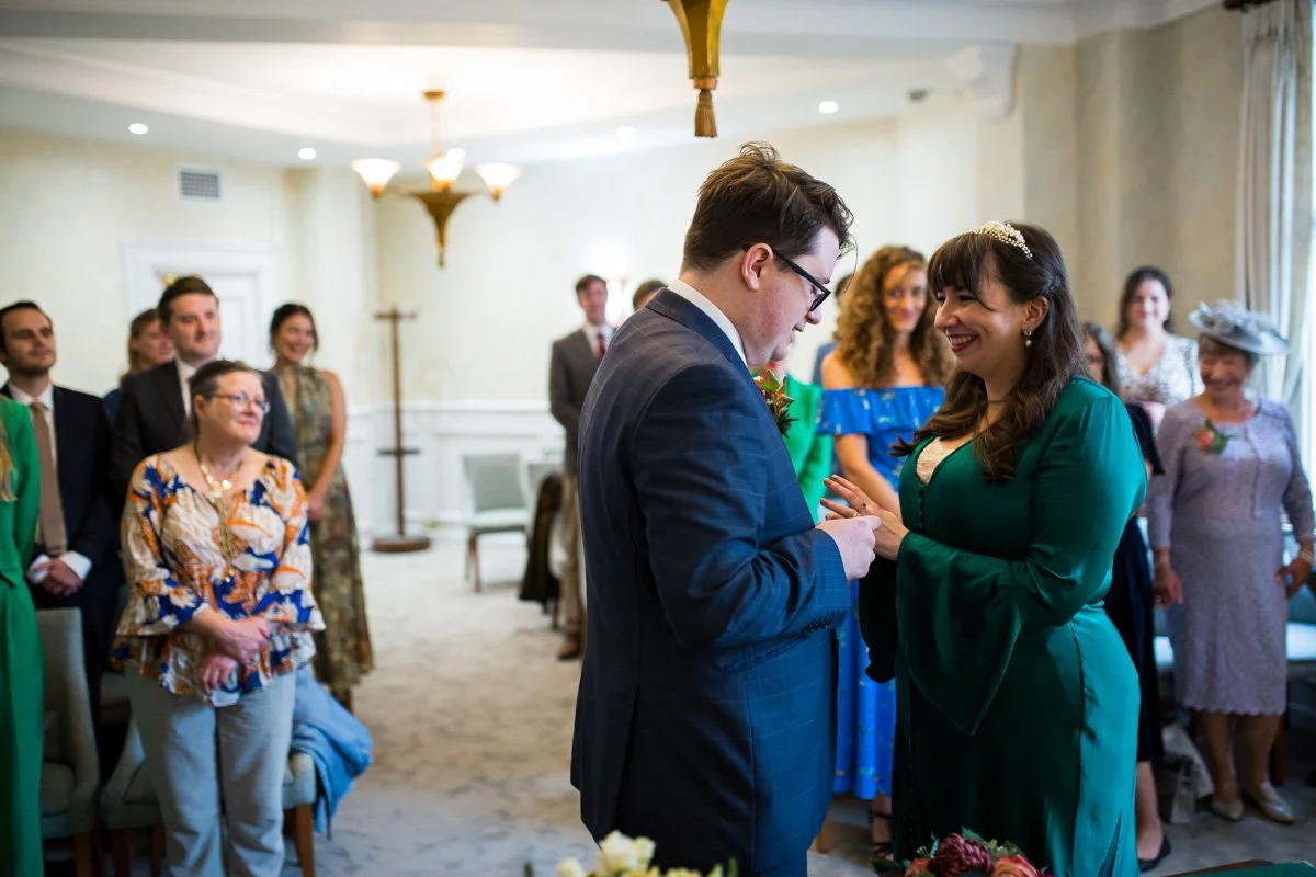 A woman in a green dress is smiling and placing a ring on the finger of a man in a gray suit during a wedding ceremony, with guests watching in a softly lit room.
