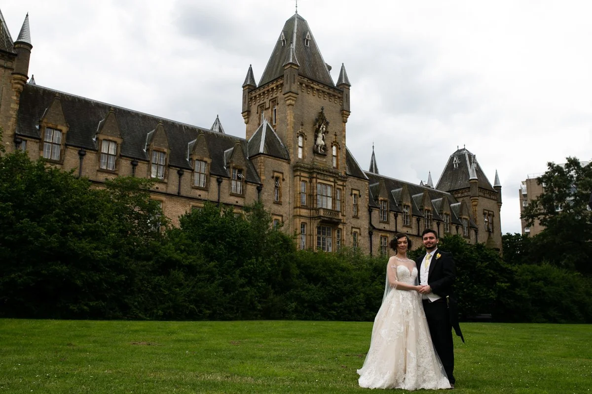 A bride and groom are standing hand in hand on a green lawn in front of an old castle-like building with turrets and stone walls. The bride wears a white wedding dress and veil, and the groom wears a black suit with a white shirt and yellow tie. Over