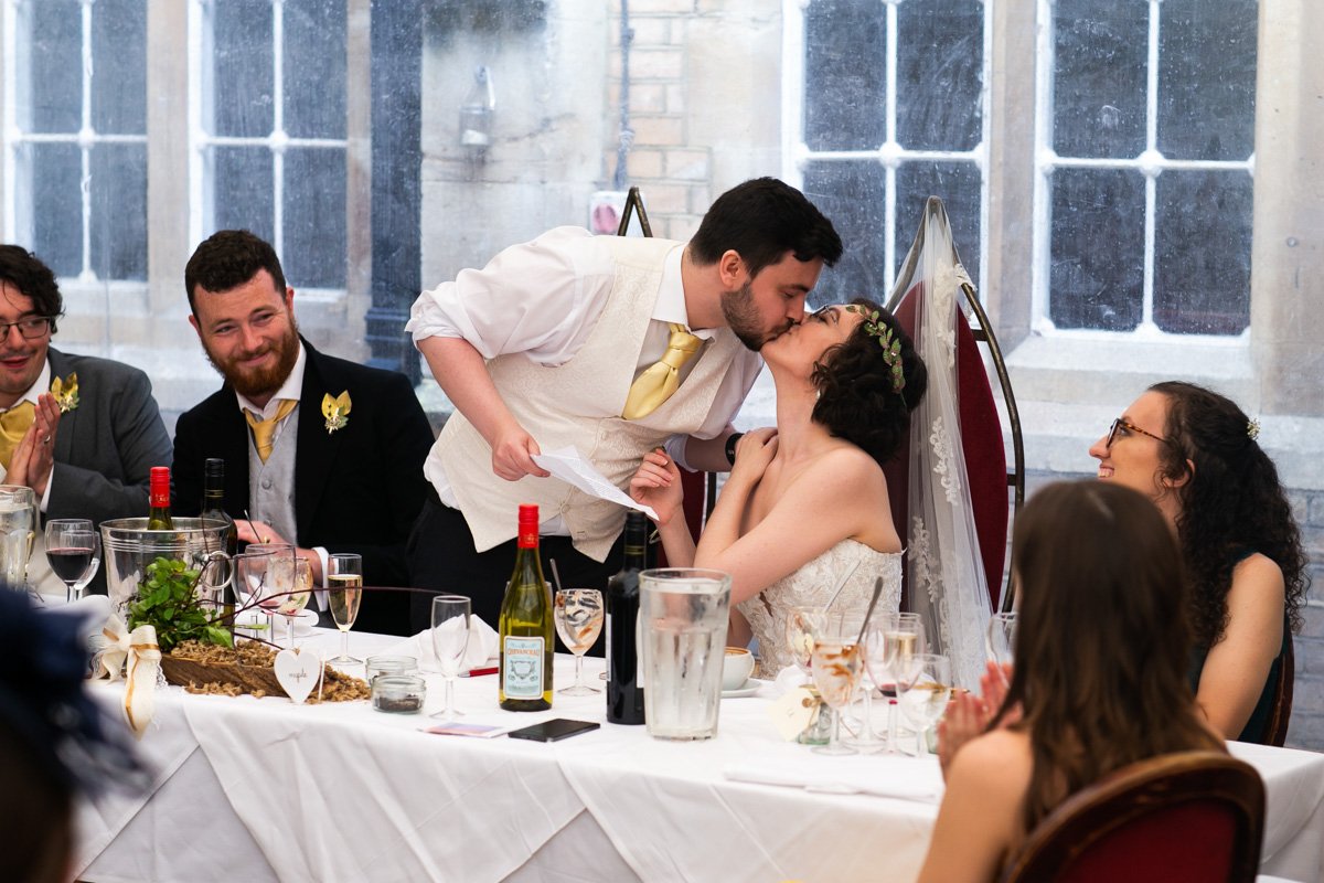 A wedding reception with a groom kissing the bride, seated at a decorated table, as guests clap and smile around them.