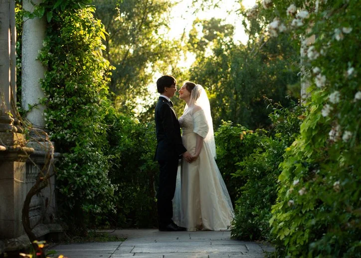 A bride and groom standing close together, holding hands, in a lush garden during sunset, sharing a moment before their wedding.