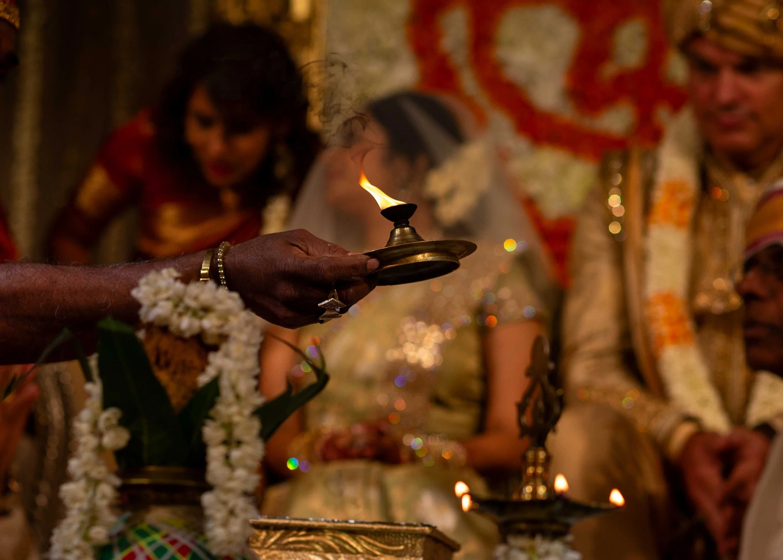 Close-up of a person holding an oil lamp with a flame during a traditional ceremony, surrounded by other participants dressed in colorful traditional attire.