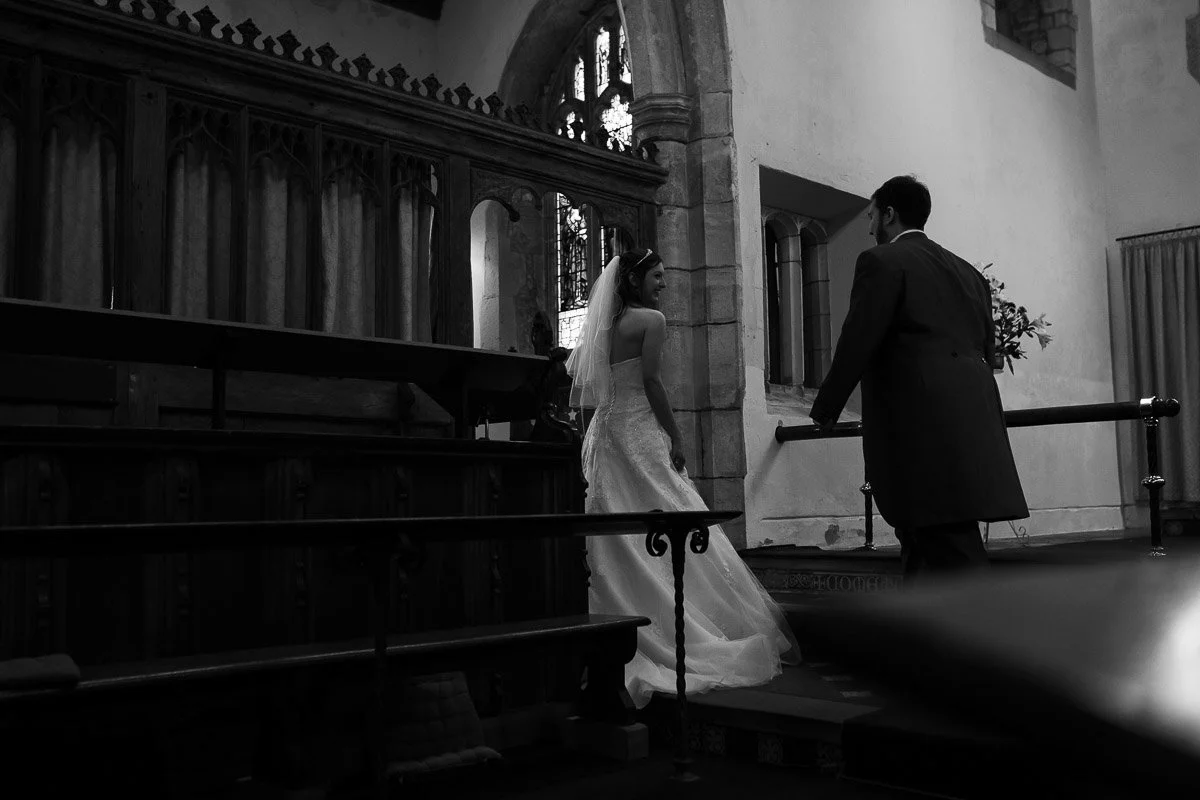 A bride and groom standing inside a church, facing each other, with stained glass windows and church architecture visible in the background.