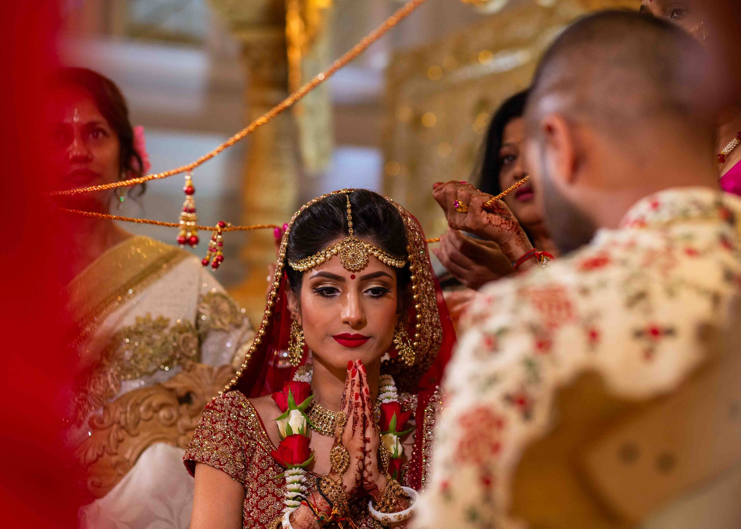 An Indian bride dressed in traditional red and gold wedding attire, with jewelry and henna on hands, sits with hands in a prayer gesture, while a groom and women participate in a religious ceremony.
