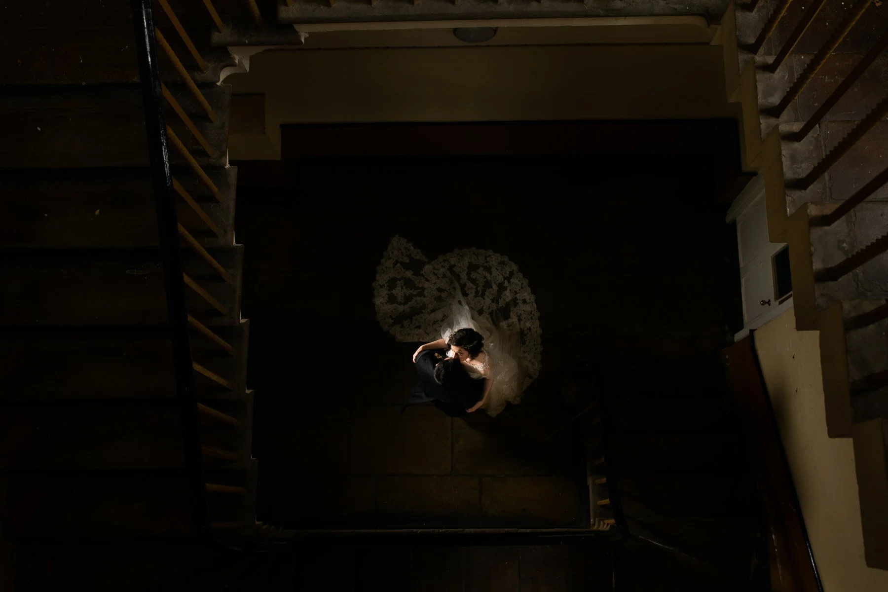 A wedding bride and groom embracing on a dark staircase, viewed from above, with soft lighting highlighting her white dress and veil.
