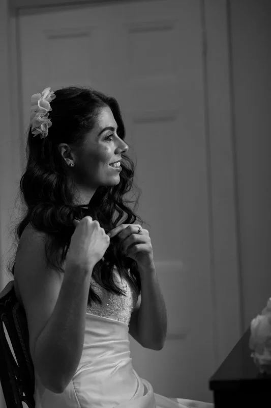 A woman with long, wavy hair and a flower in her hair, sitting at a table and smiling, in black and white.