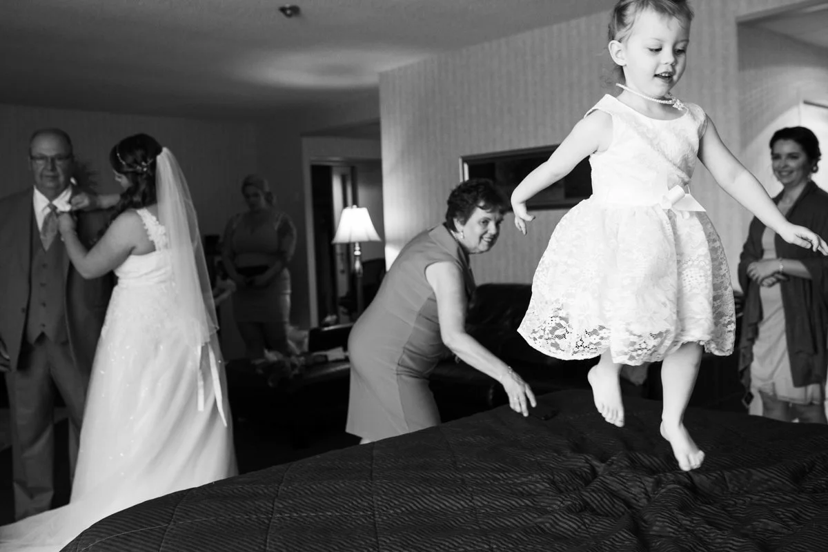 A young girl in a lace dress jumps on a bed with a big smile. In the background, a bride in a wedding gown dances with an older man, and several women are standing and watching in a warmly lit room.