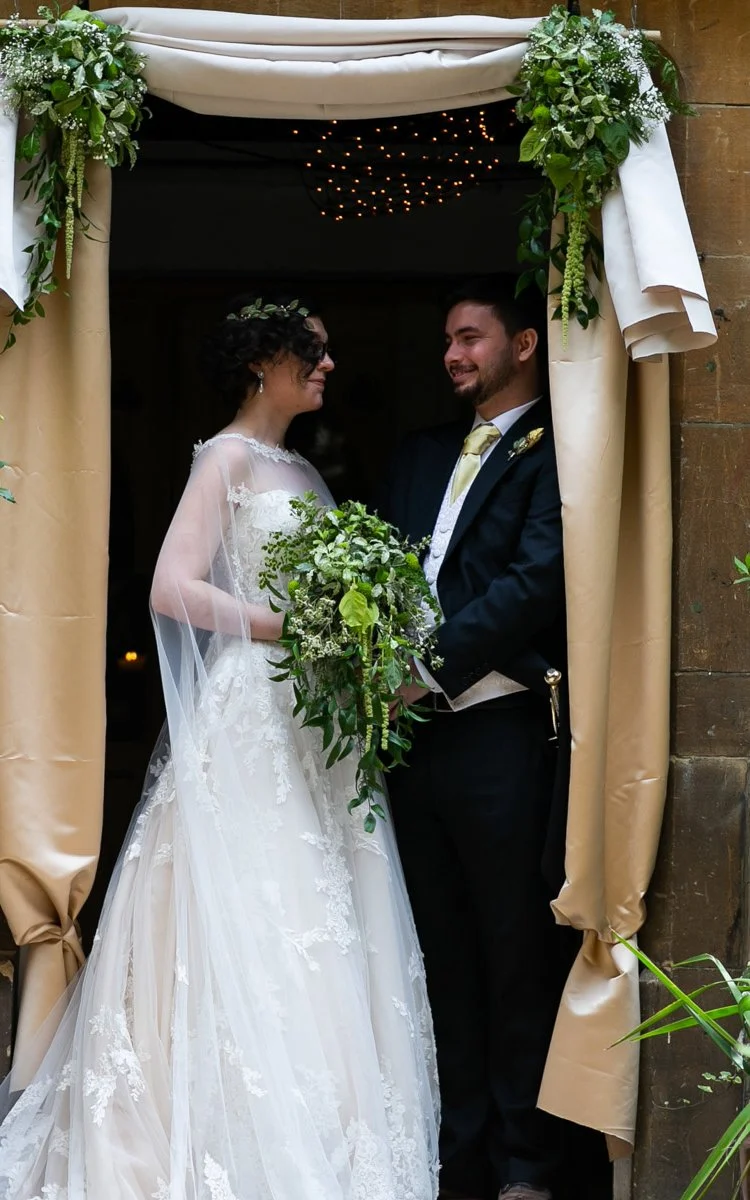 A bride and groom standing together under a decorated arch, holding a bouquet of flowers, smiling at each other during a wedding ceremony.