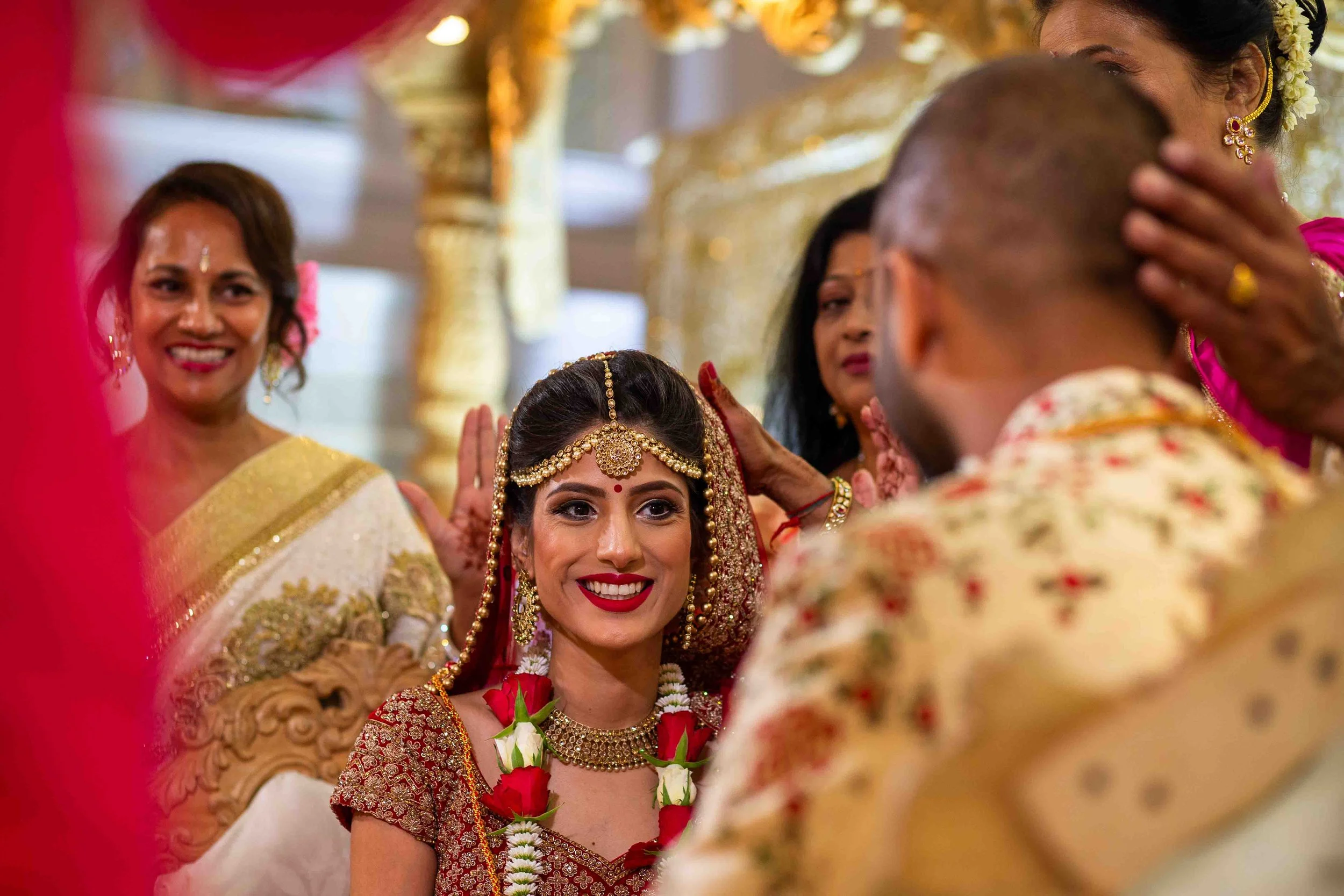 An Indian bride with jewelry and a floral garland smiling as she is blessed by a groom during a traditional ceremony, surrounded by women in colorful sarees.