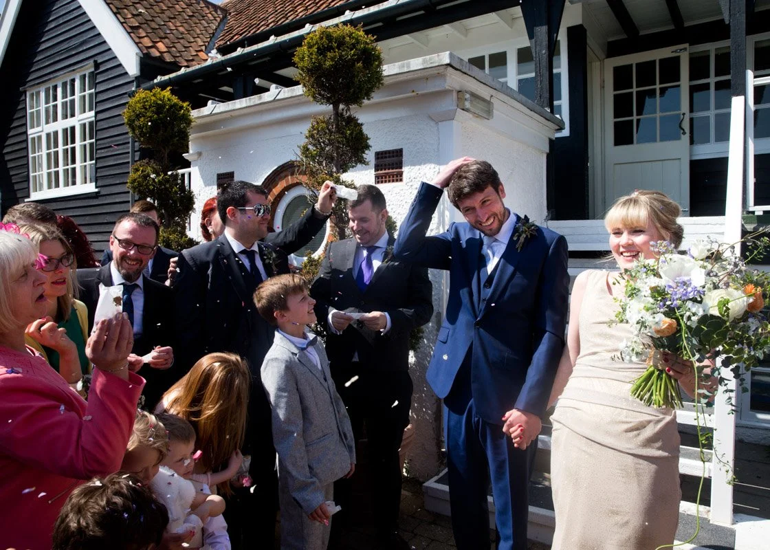 A cheerful bride and groom holding hands outside a house, surrounded by friends and family celebrating a wedding, with confetti in the air.