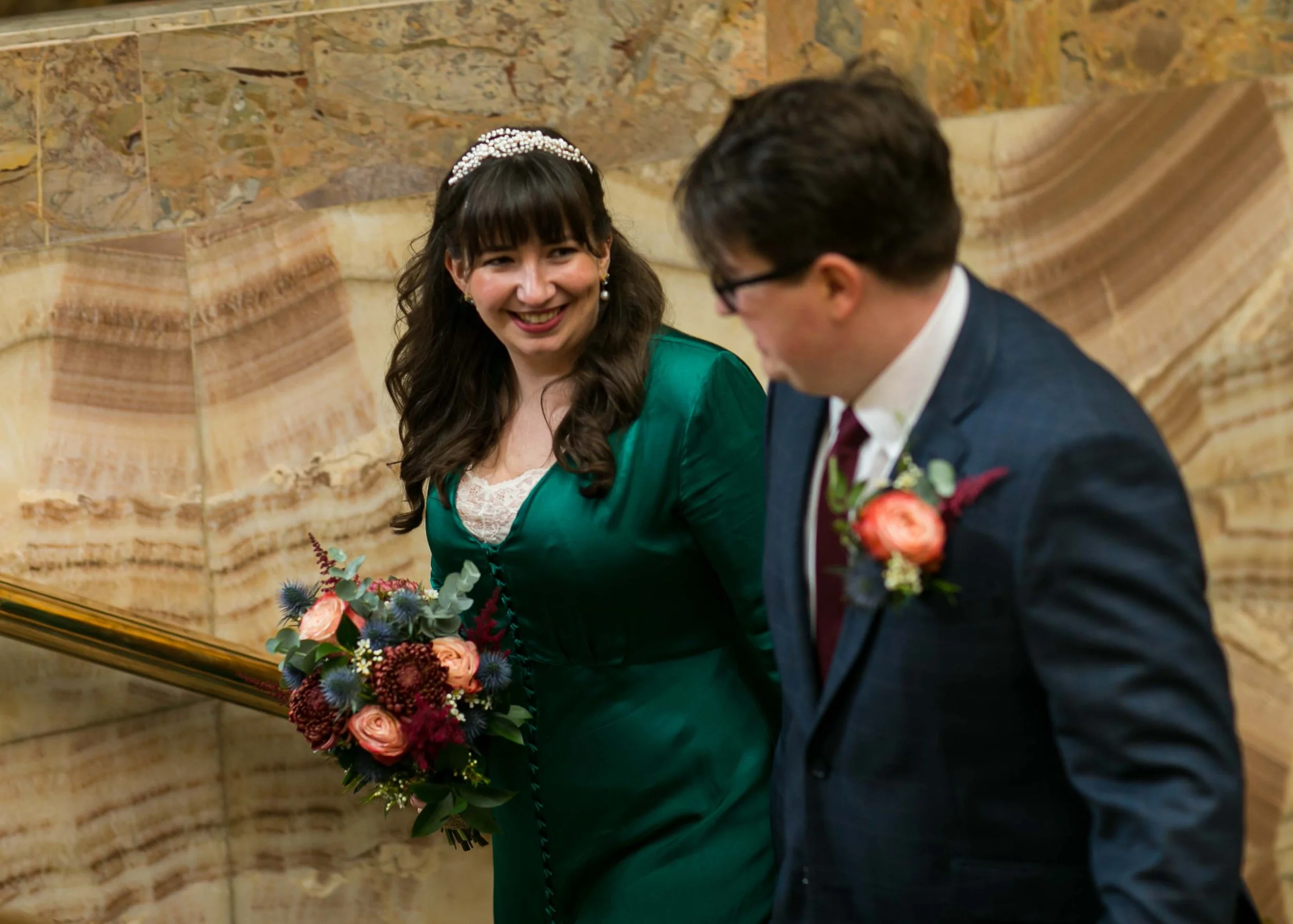 A woman and a man are sitting together at a piano, smiling at each other. The woman is wearing a green jacket and a pearl headband, holding a colorful bouquet of flowers. The man is dressed in a suit with glasses and a boutonniere. They are in front 