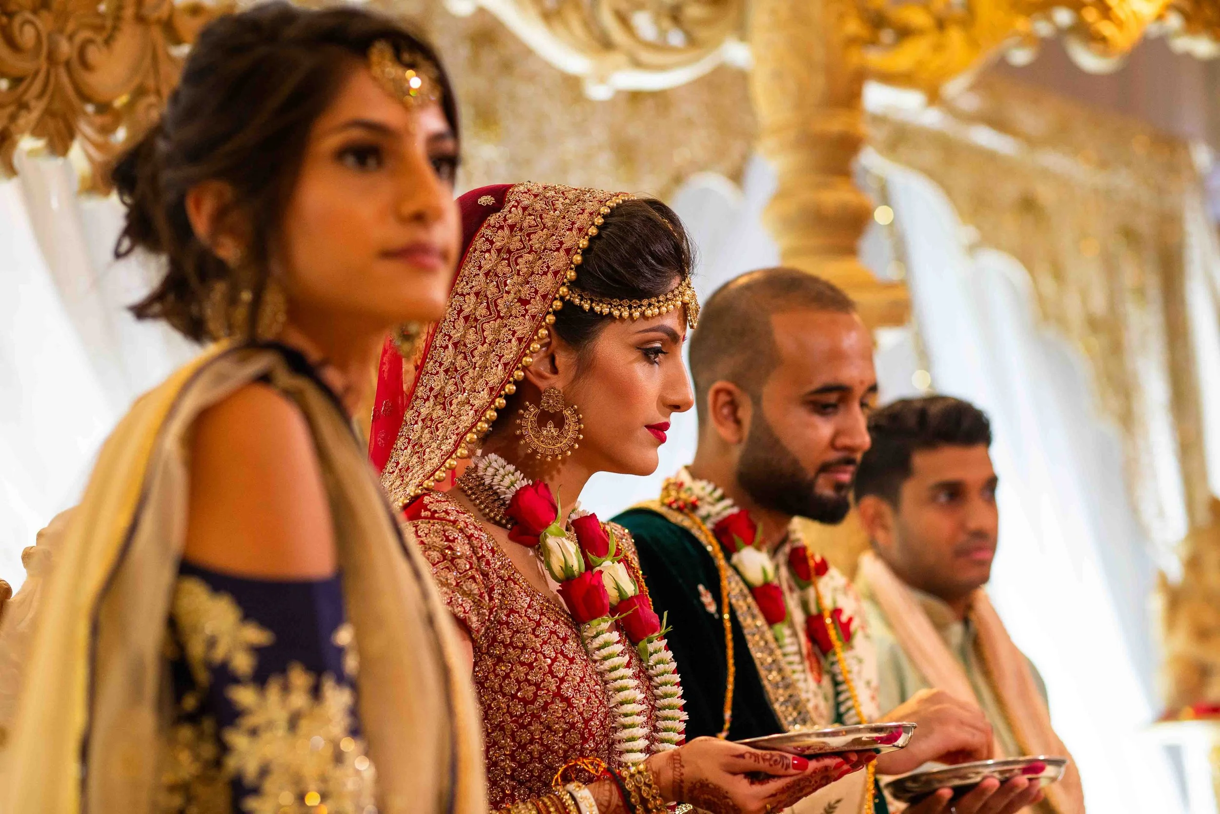 Indian bride with jewelry and floral garlands standing with her family during a wedding ceremony.