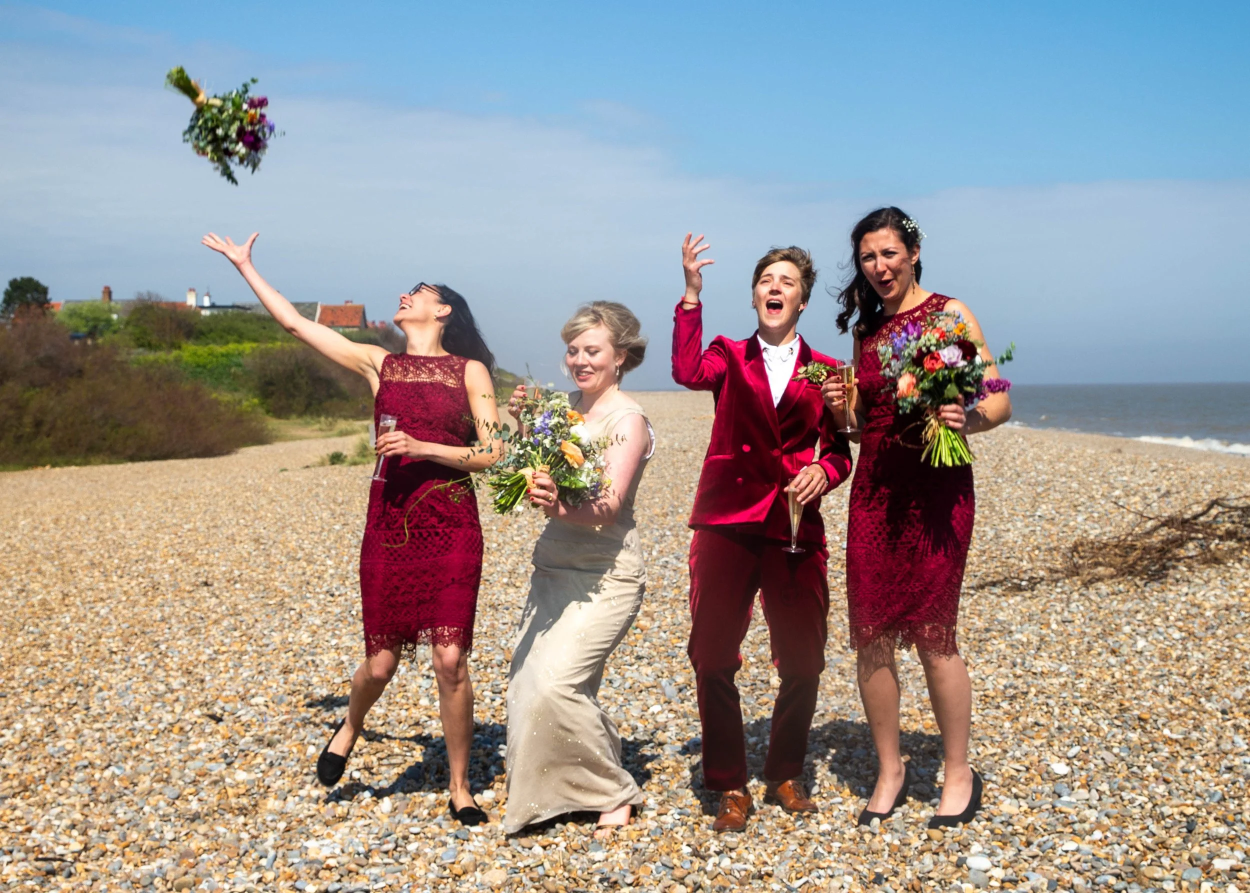 A group of five women on a pebble beach, dressed in elegant semi-formal attire, celebrating with bouquets and glasses of champagne, as one woman throws a flower bouquet into the air.