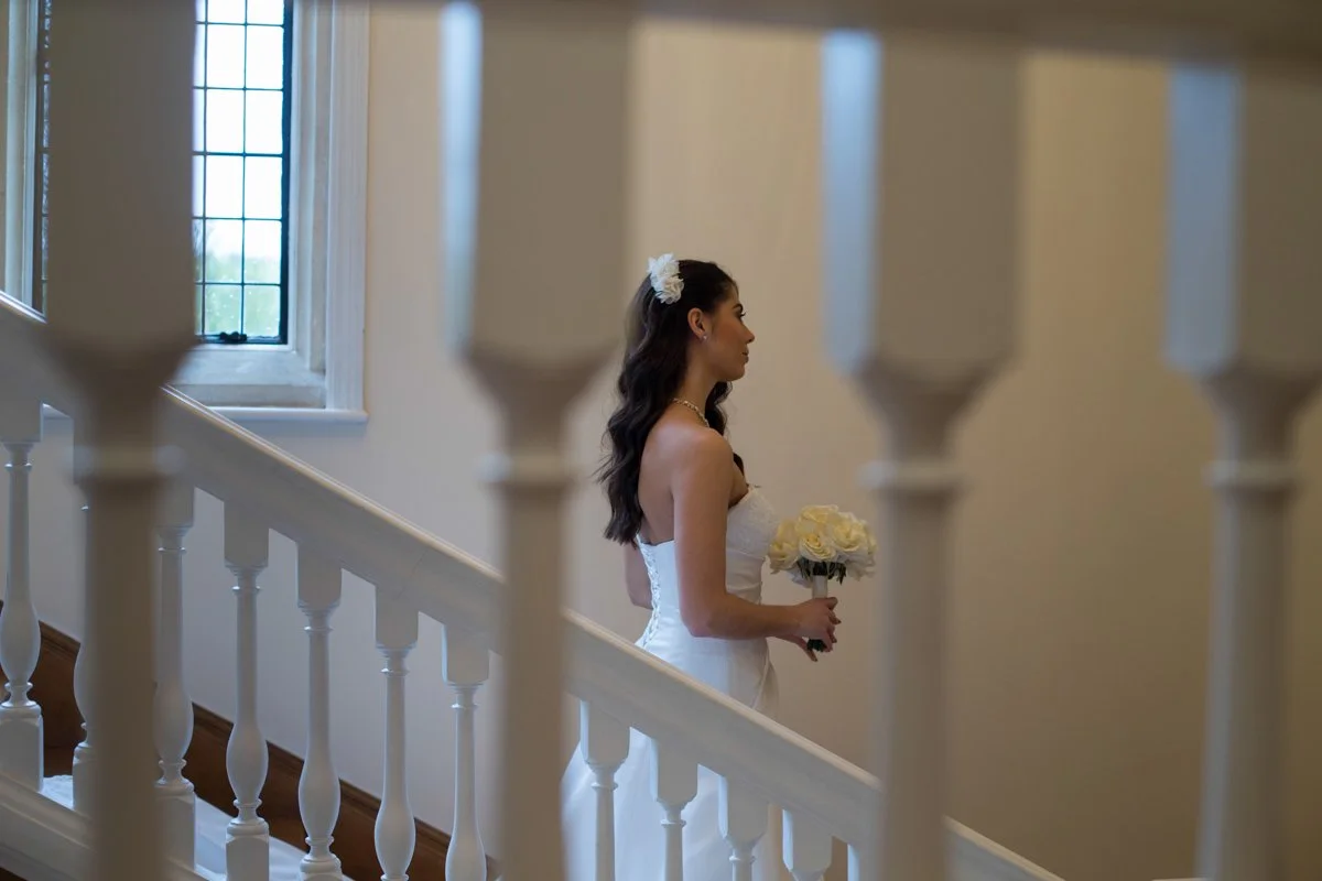 A bride in a white wedding dress holding a bouquet of white roses, standing on a staircase near large windows.