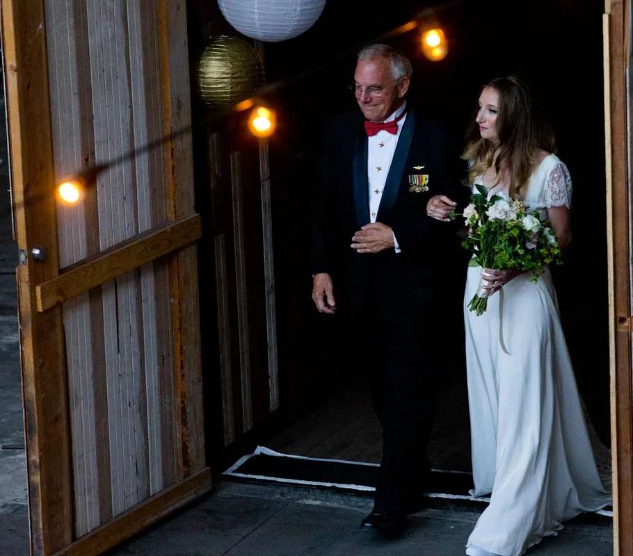 A bride in a white wedding dress holding a bouquet walks down the aisle supported by an older man in a tuxedo with medals. They are entering a decorated venue with string lights and paper lanterns.