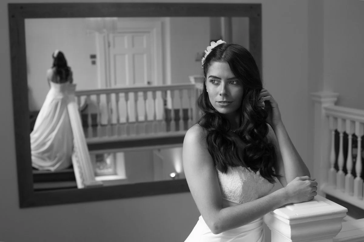 A young woman in a wedding dress with long dark hair and a flower crown, looking thoughtfully while touching her hair, standing in front of a large mirror in a house interior with a staircase.