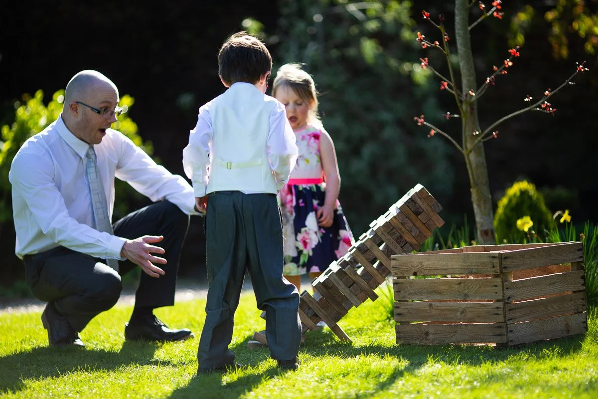 A man in a white shirt and tie is crouched down talking to two children outdoors on a sunny day, with a playground toy made of wooden pallets in front of them and a small tree with red flowers nearby.