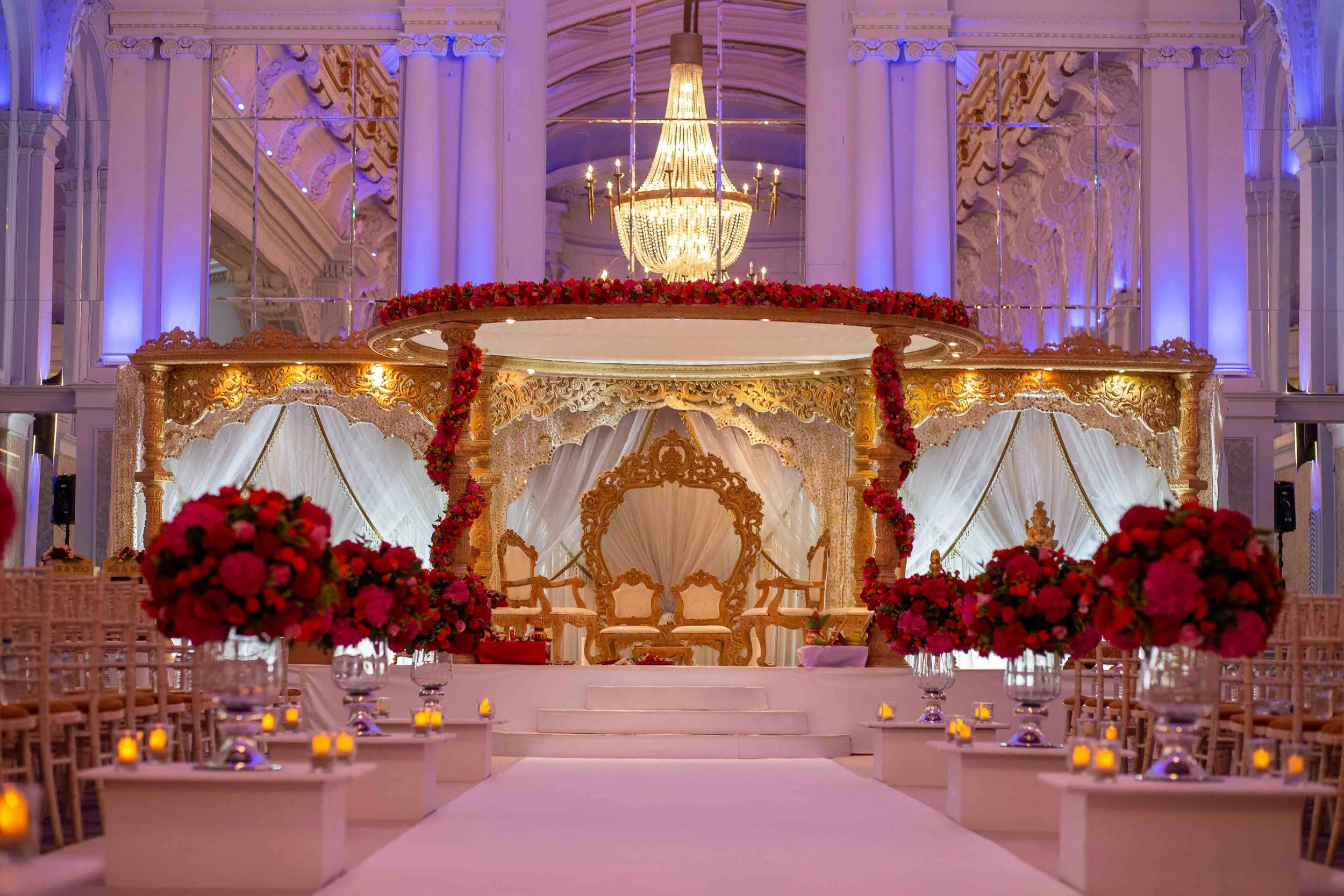 Elegant wedding stage with ornate golden furniture, surrounded by pink and red floral arrangements, in a grand hall with purple lighting and a large chandelier.