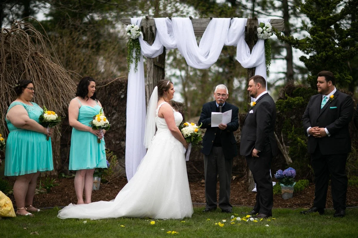 Outdoor wedding ceremony with bride and groom facing each other under a decorated wooden arch, with bridesmaids and groomsmen standing beside them, the officiant reading vows or a speech, on a grassy area surrounded by trees.