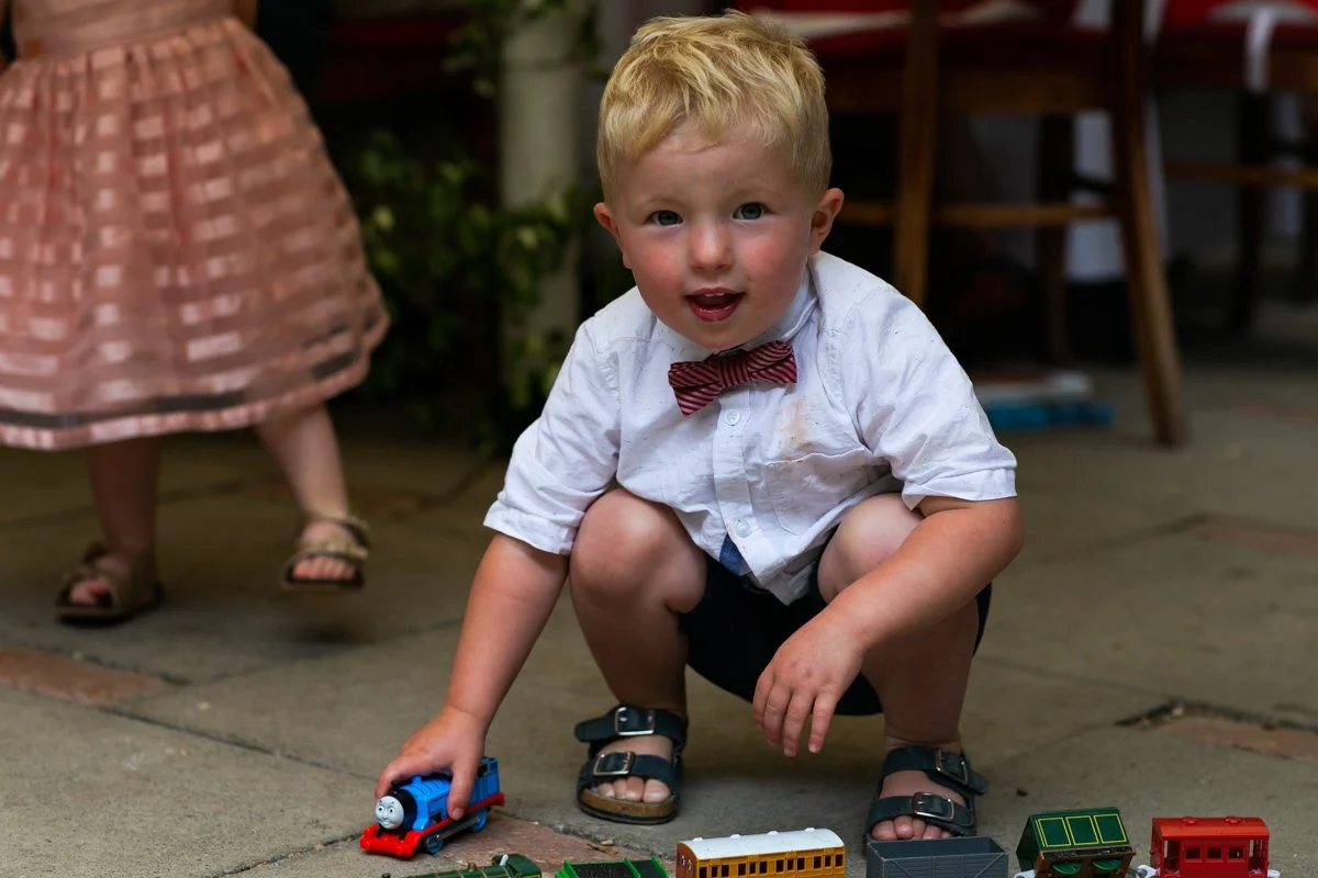 A young boy with blonde hair, wearing a white shirt, red bow tie, and black shorts, squatting on the patio playing with a blue toy train. An adult woman in a pink patterned skirt and sandals stands nearby.