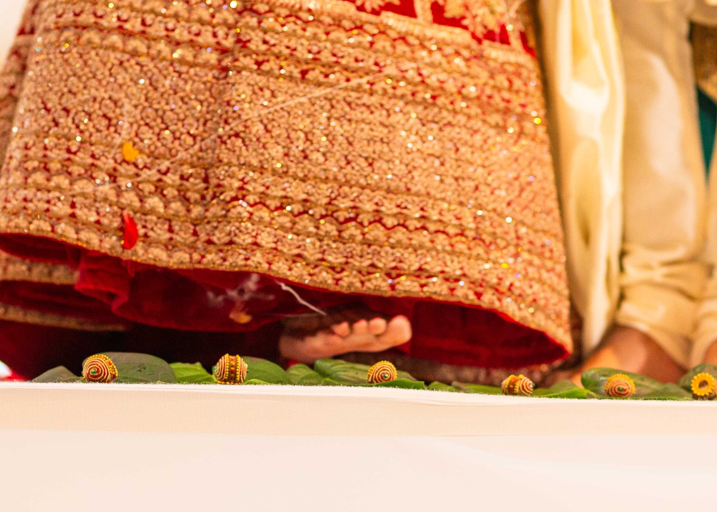 Close-up of a person in traditional Indian attire, possibly during a religious or cultural ceremony, with their hand placed on a decorated surface. The surface is adorned with green leaves and small ornamental objects, including miniature family symb