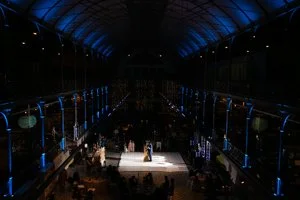 A fashion runway show with models walking down the catwalk in a dark, illuminated venue with blue and white lighting.
