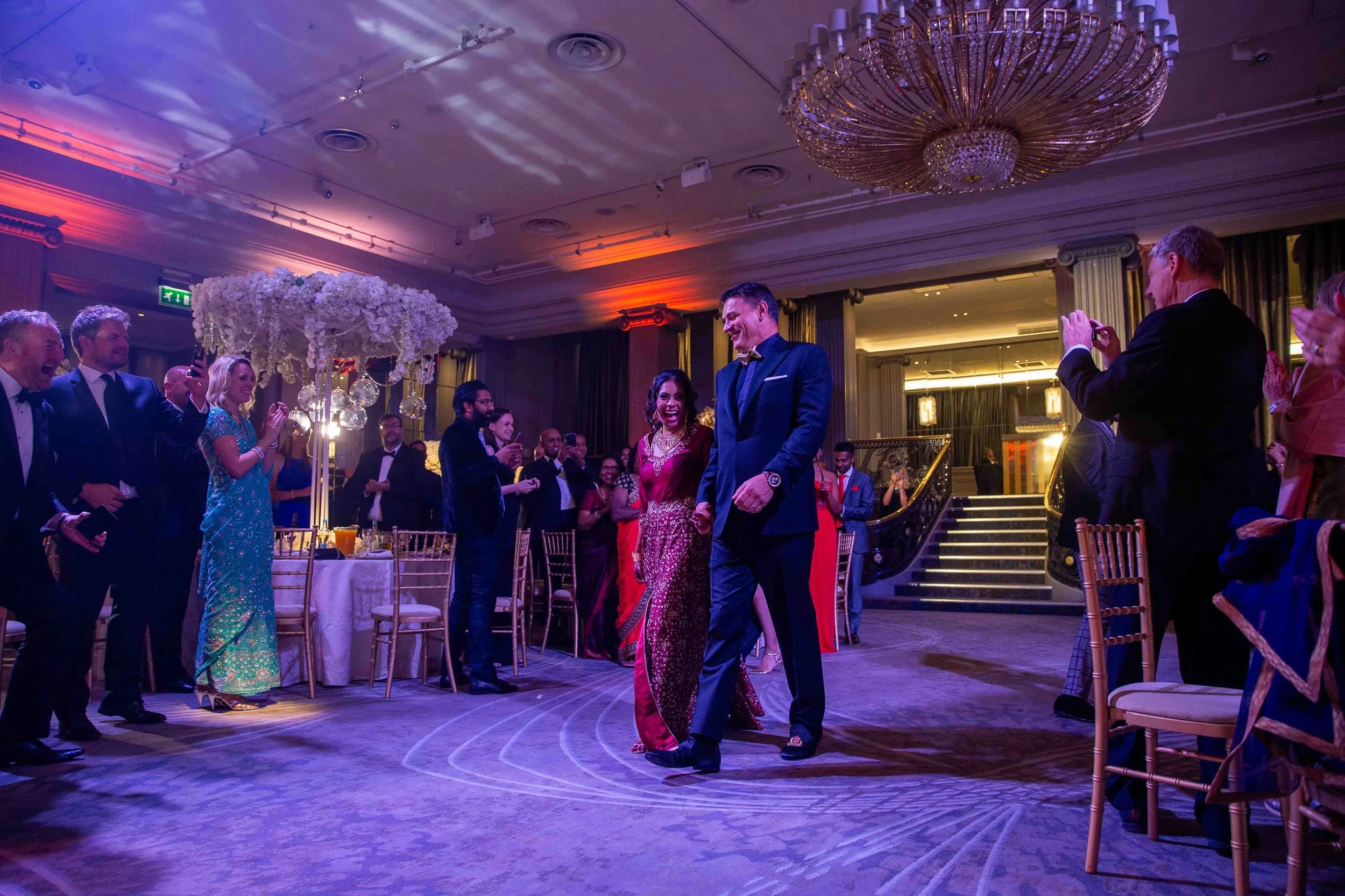 A group of people at an elegant event or wedding reception, with a woman in a traditional red and gold dress and a man in a dark tuxedo walking together on the dance floor, surrounded by guests clapping and taking photos, indoors with a chandelier an