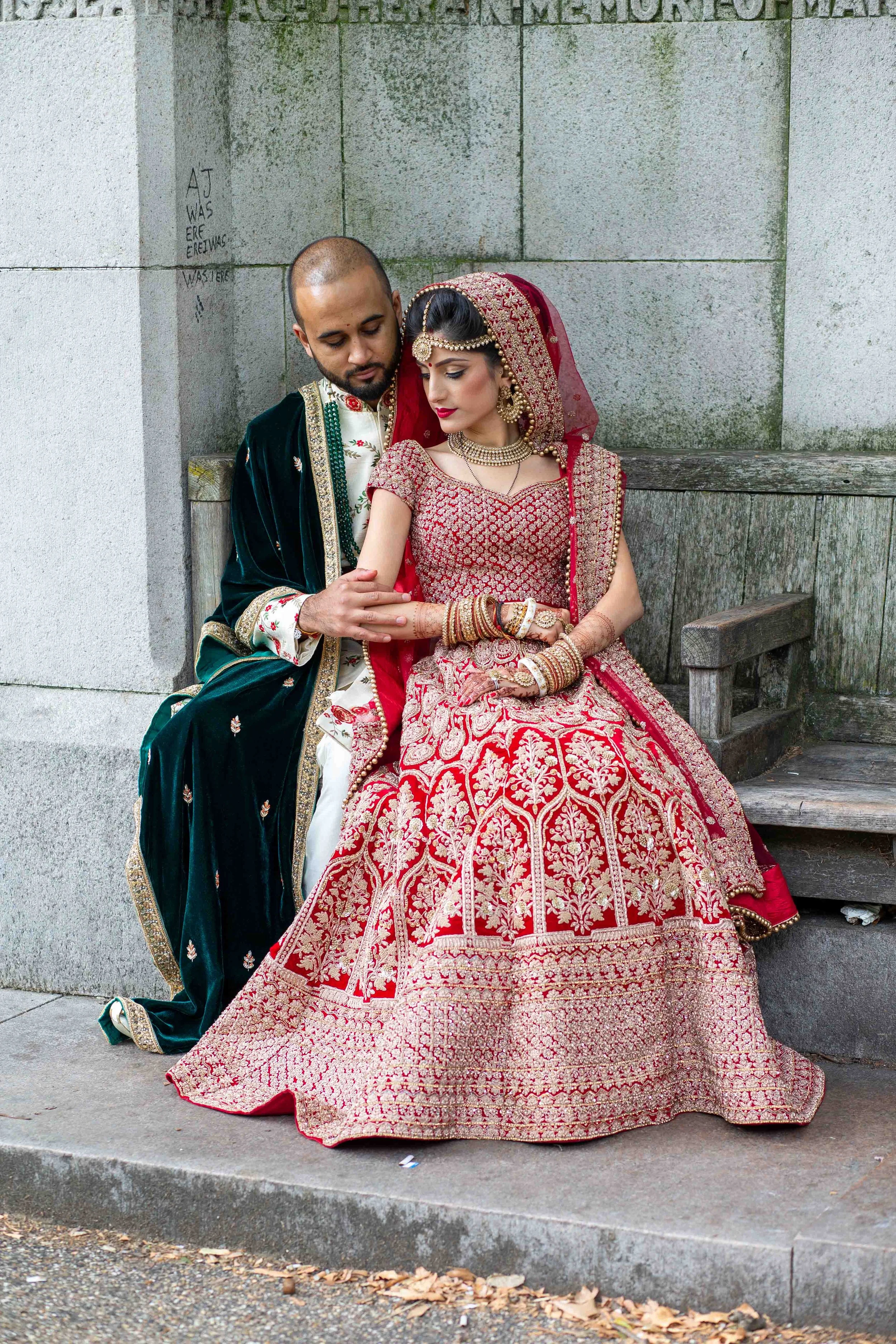 An Indian bride and groom dressed in traditional wedding attire sitting on a park bench.