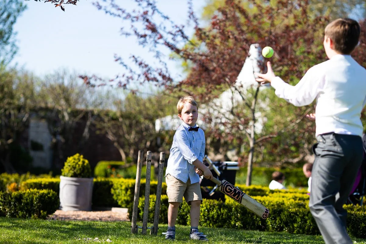 Two young boys playing cricket outdoors on a sunny day, one holding a cricket bat and the other throwing a tennis ball.