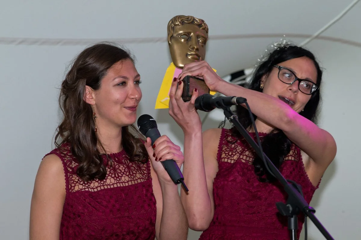 Two women in matching maroon dresses are on stage during an award ceremony. One woman is holding a microphone, and the other is holding an award resembling a golden face sculpture. The woman with glasses appears surprised or emotional.