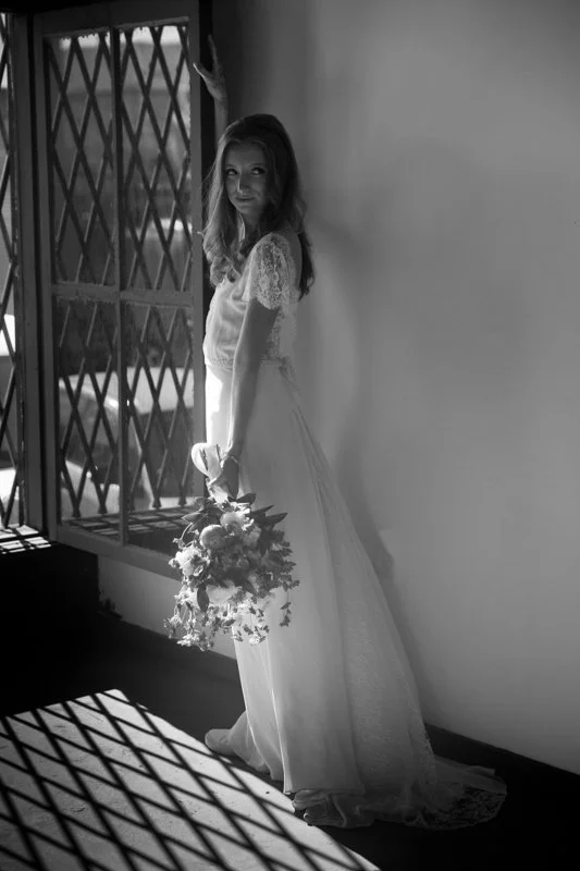 A bride in a wedding dress holding a bouquet, standing near a window with sunlight casting shadows on the floor.