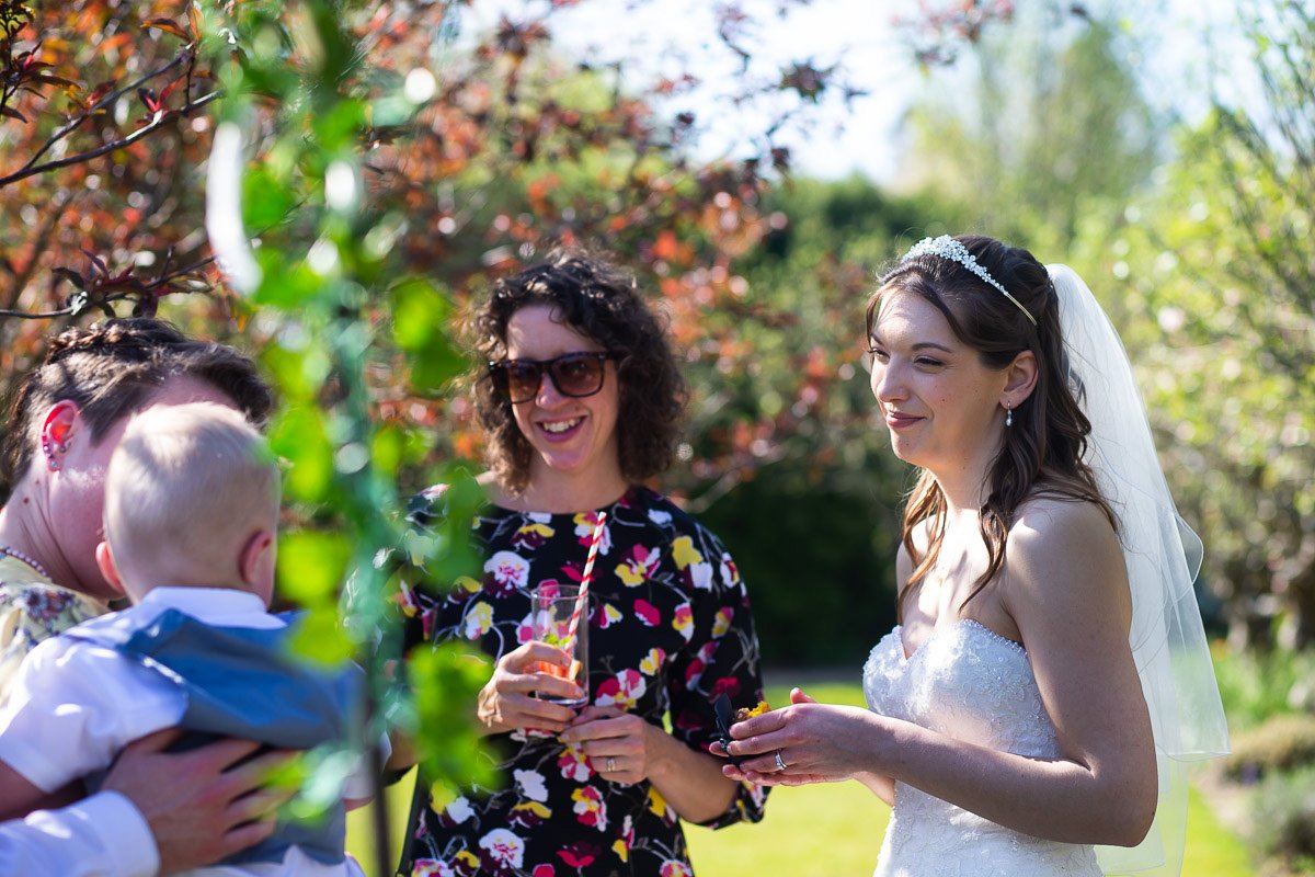 Bride in a wedding dress and veil talking to a woman and a baby outdoors with trees and greenery in the background.
