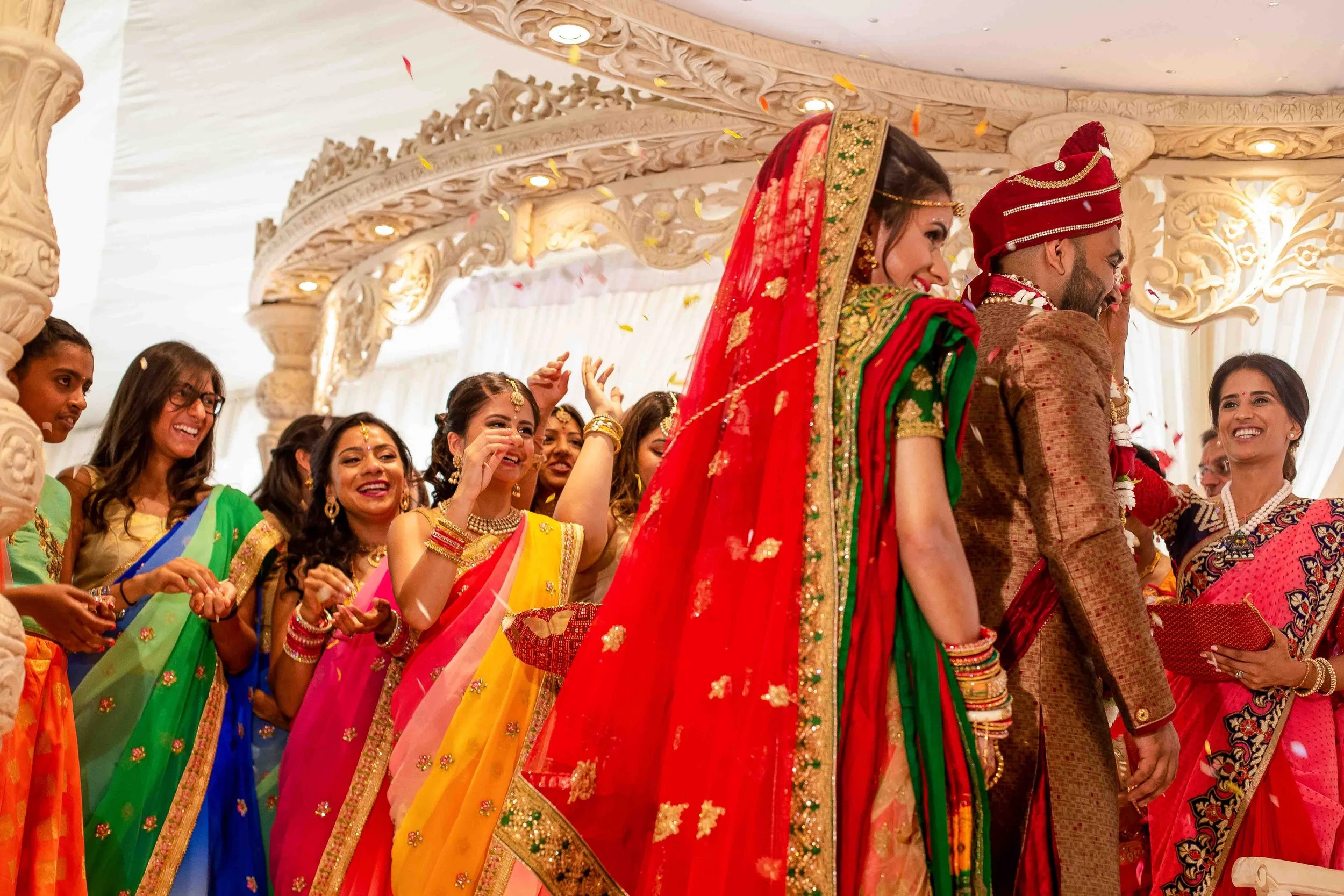 Indian bride and groom at a wedding ceremony, surrounded by women in colorful sarees, celebrating and smiling under ornate decorations.