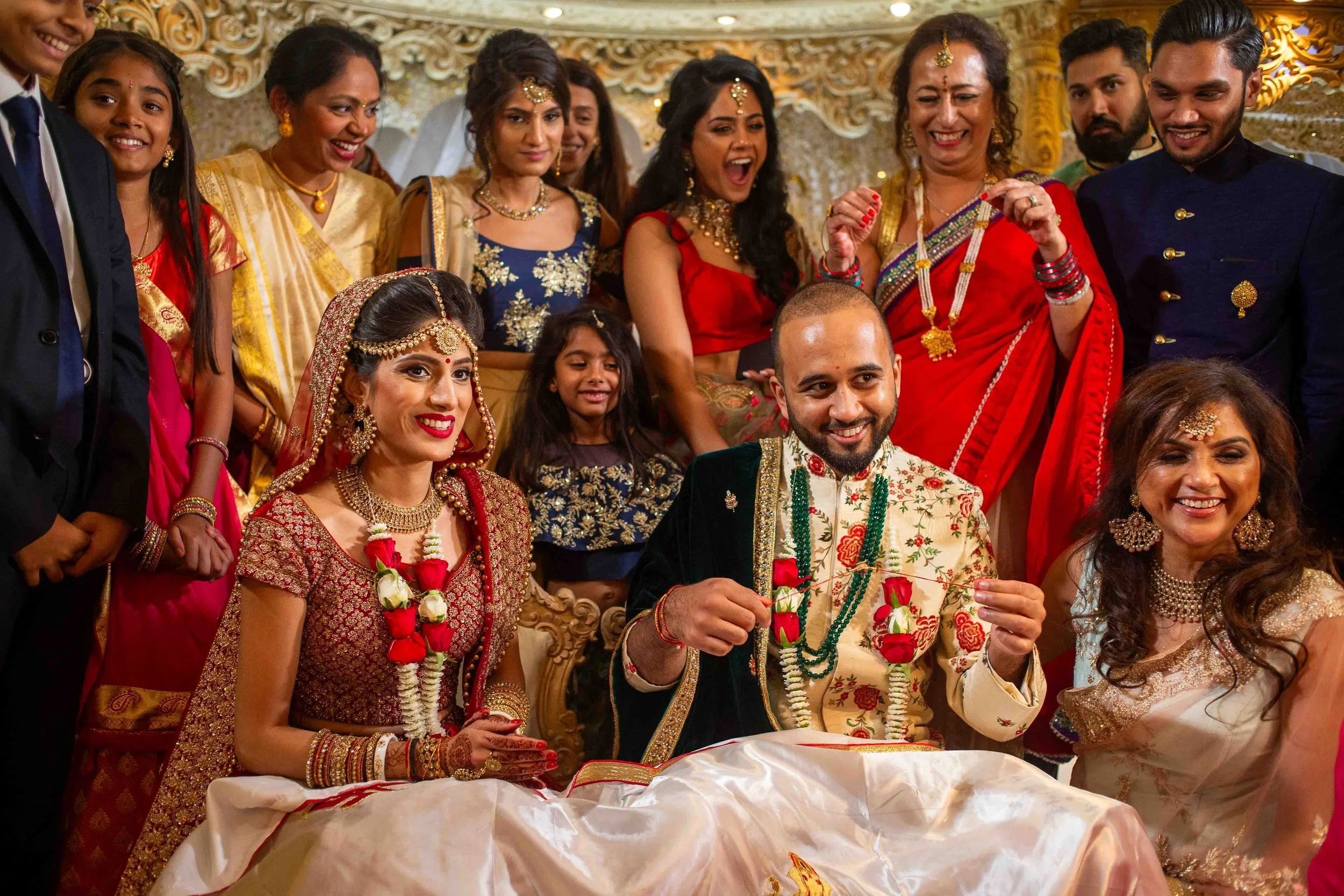 A traditional Indian wedding ceremony with the bride and groom seated, surrounded by family and friends wearing colorful ethnic attire. The bride is dressed in a red and gold sari, and the groom is in a cream and gold embroidered outfit. Everyone is 