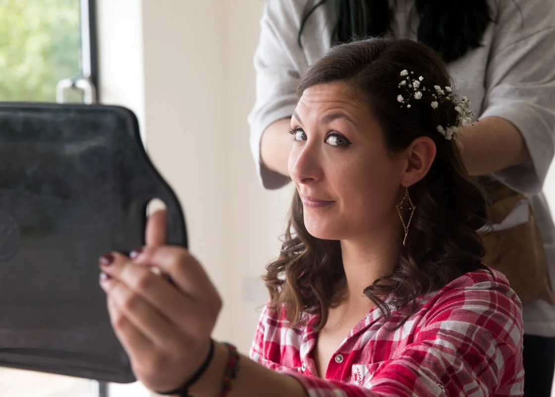 A woman with dark curly hair decorated with white baby's breath flowers is taking a selfie with her phone while another person styles her hair behind her.